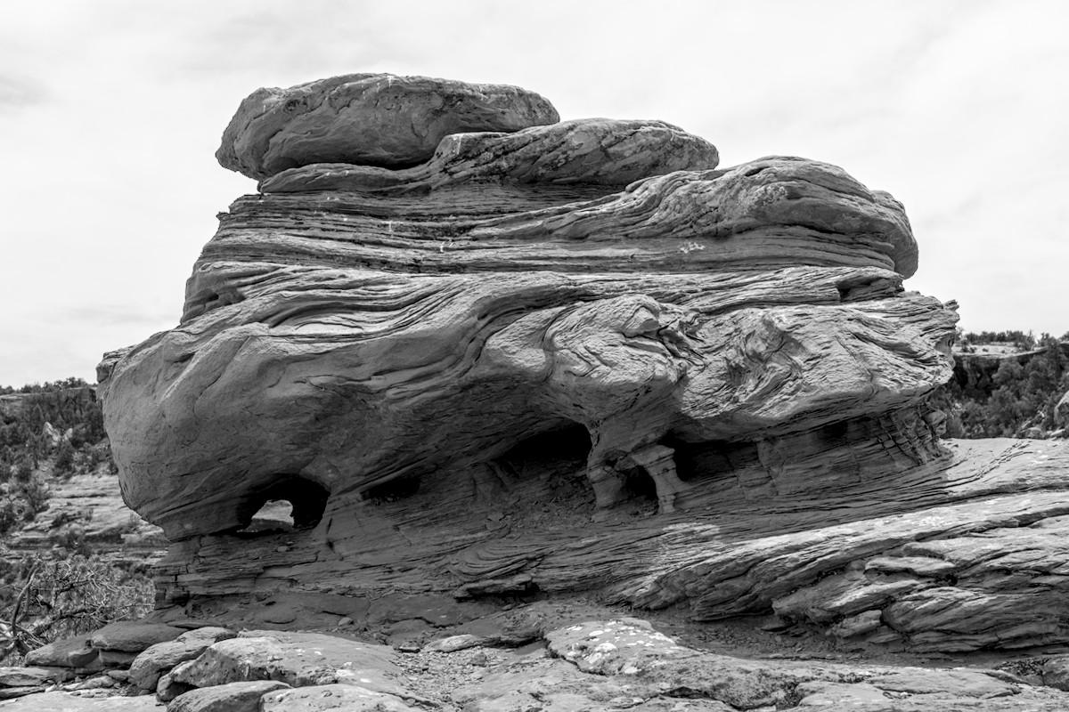 A black and white landscape photo of a highly eroded and sculpted sandstone boulder. It has many horizontal layers stacked on top of one another. The bottom, and widest layer has many curves in it. The base is undercut and there are a couple of very small <br />arches. The whole thing is the size of a large house.