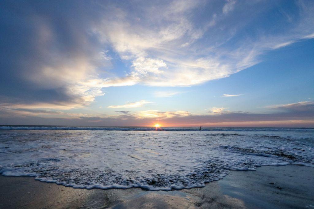 An ocean wave gently spreads across the shore at sunset—the sun is just touching the horizon, clouds above mimicking the shape of the ocean wave. A person stands in the water in the distance with arms outstretched.