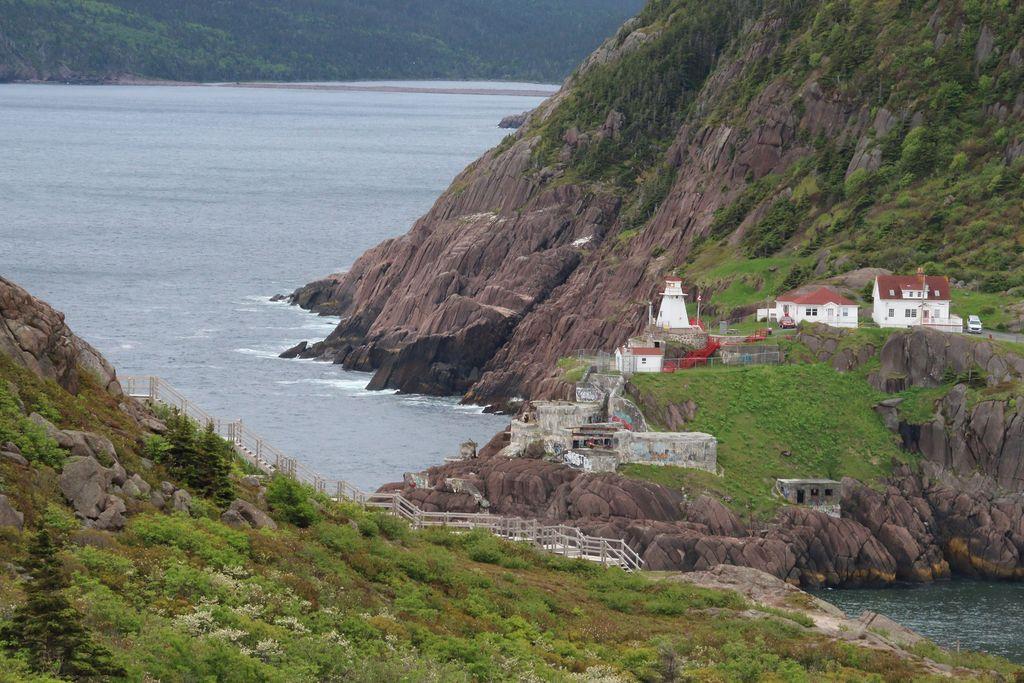 Photo of a cove on the cost of Newfoundland, at Signal Hill. Rocks jut out of the water, with a wooden staircase leading down one rocky hill, and a lighthouse and some historic buildings planted on the rocky terrain of another.