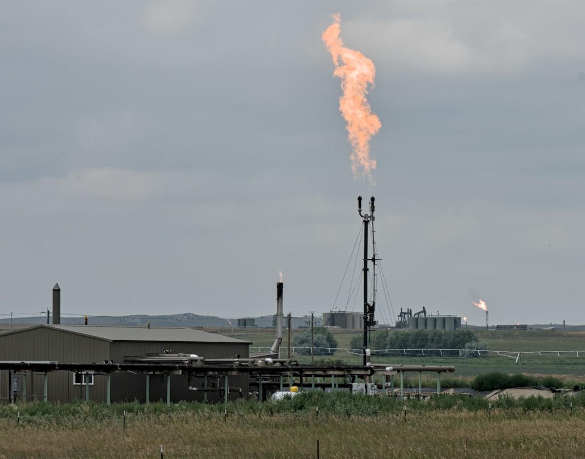 A color landscape photo showing a single story brown colored building in the foreground on the left and extending out toward the center of the photo from the building is a network of pipes. Three moderately tall stacks are seen, one behind the building, one just to the right of the building, and another taller one with a forked end further to the right. The last two spout flames with the flames being very long from the stack furthest to the right. In the background further to the right is another building with stacks and flames. If the viewer looks closely, very tiny dots can be seen further out in the photo indicating yet more gas flares. A color landscape photo showing a single story brown colored building in the foreground on the left and extending out toward the center of the photo from the building is a network of pipes. Three moderately tall stacks are seen, one behind the building, one just to the right of the building, and another taller one with a forked end further to the right. The last two spout flames with the flames being very long from the stack furthest to the right. In the background further to the right is another building with stacks and flames. If the viewer looks closely, very tiny dots can be seen further out in the photo indicating yet more gas flares.