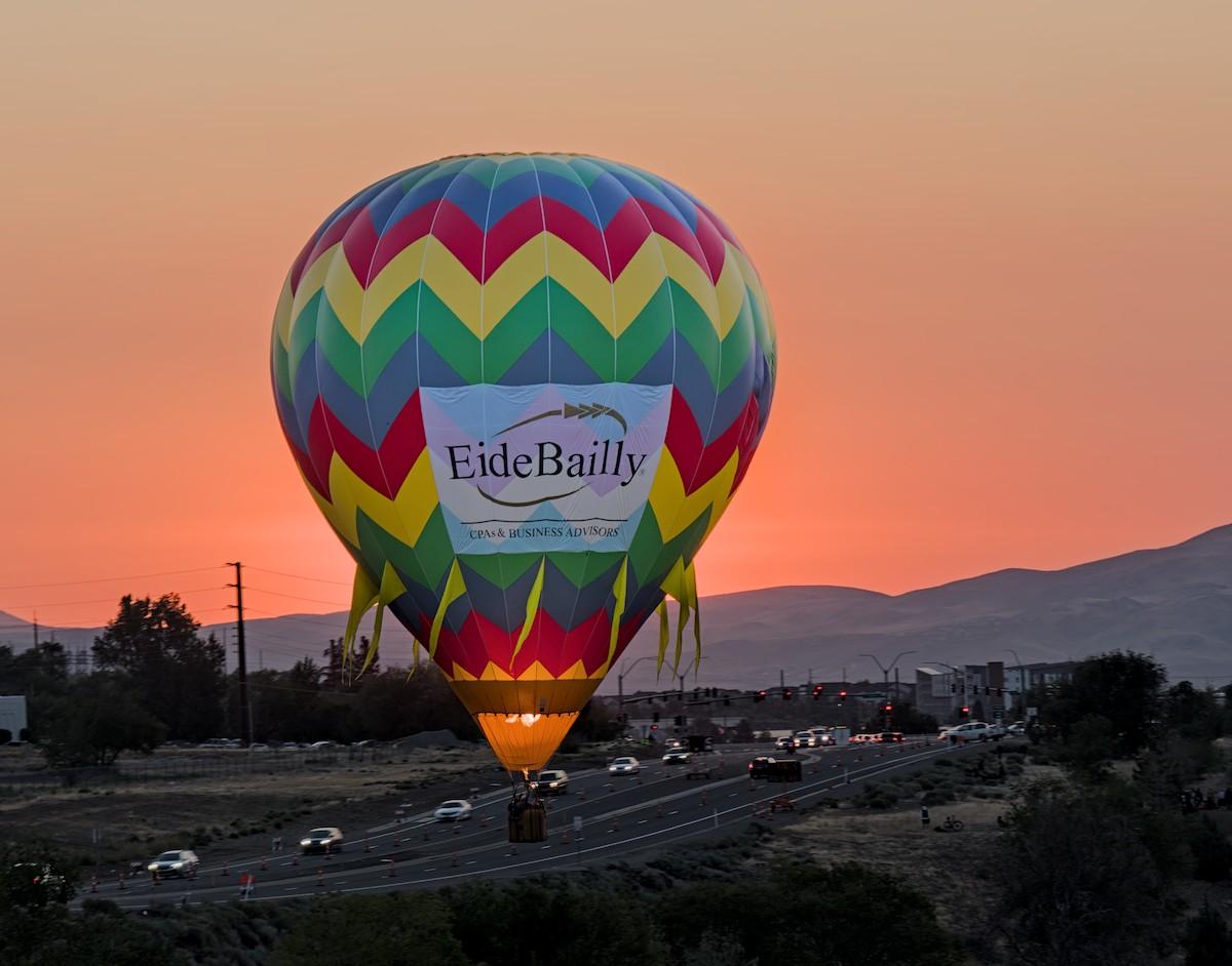 A color landscape photo of a hot air balloon with a orange sunrise behind it. The balloon hangs low over a highway. The balloon has zig-zag stripes of blue, red, yellow, and green going around it. Sadly, there's a big white advertisement banner facing the camera ruining the image. The balloon also has long tassels hanging from the lower quarter part of the envelope. 