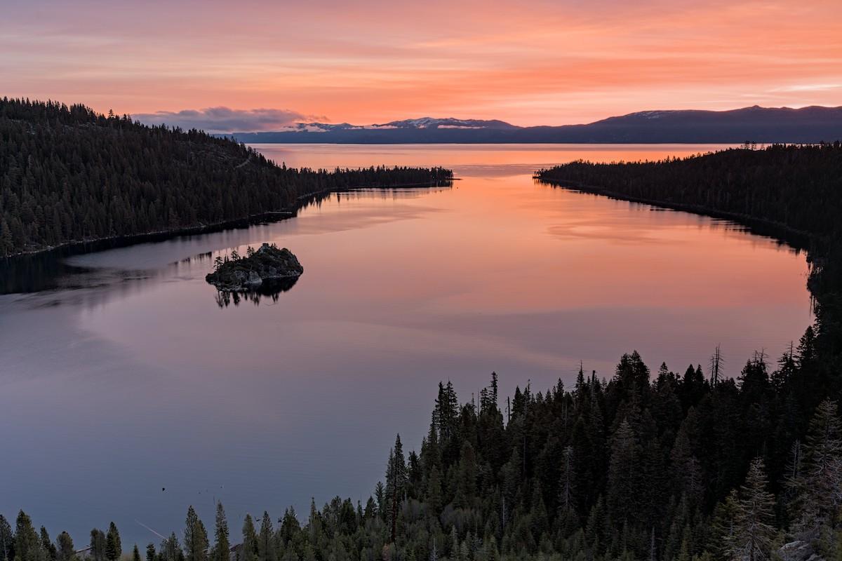 A color landscape photo showing a sky streaked with sunrise oranges and yellows over an hour-glass shaped bay in the foreground and larger lake in the background. The bay in the foreground is surrounded by steep conifer covered hills. There's a small island at center-left. There's a narrow passage above center right that leads out to the larger open lake. A line of mountains are seen in the far distance behind the opposite shore. The sunrise colors are seen reflected in the water as well. A color landscape photo showing a sky streaked with sunrise oranges and yellows over an hour-glass shaped bay in the foreground and larger lake in the background. The bay in the foreground is surrounded by steep conifer covered hills. There's a small island at center-left. There's a narrow passage above center right that leads out to the larger open lake. A line of mountains are seen in the far distance behind the opposite shore. The sunrise colors are seen reflected in the water as well.
