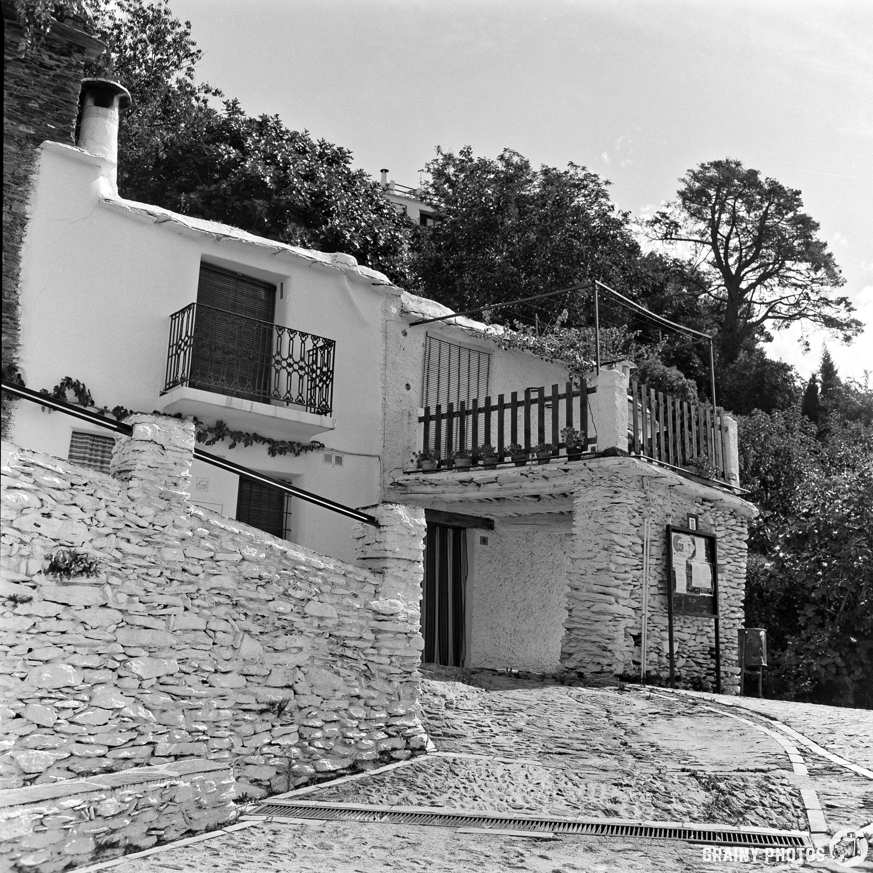 Two white stone houses with balconies and railings line a cobblestone street on a sloping terrain; this charming scene, captured in black and white on film, features trees and greenery in the background under a partly cloudy sky.