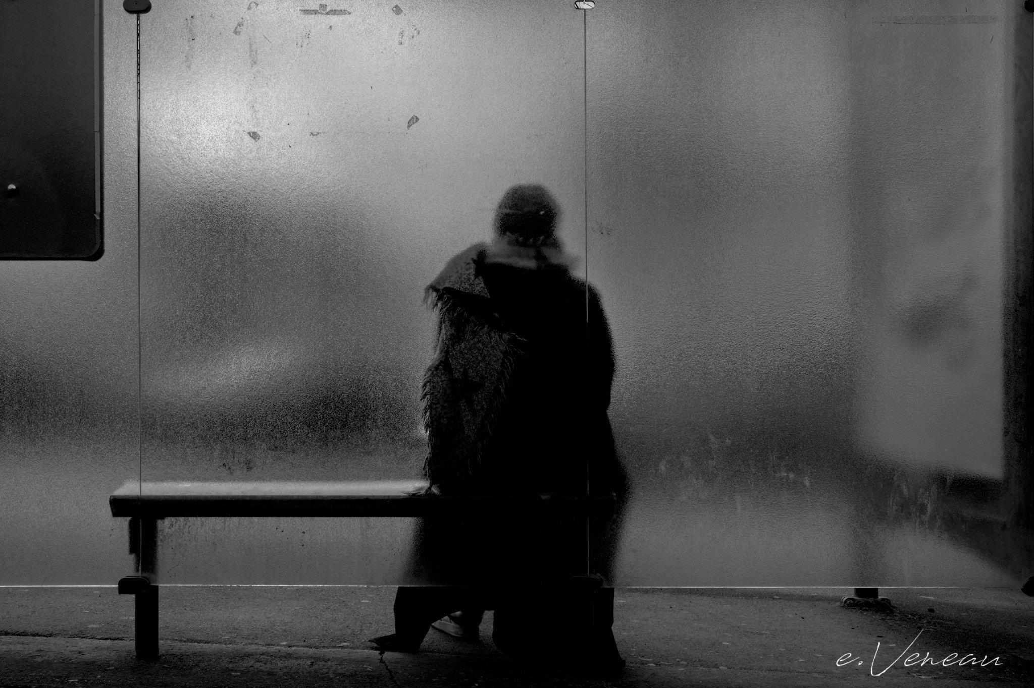 Behind the slightly opaque glass of a bus stop, a person sits waiting with his back to the bus.