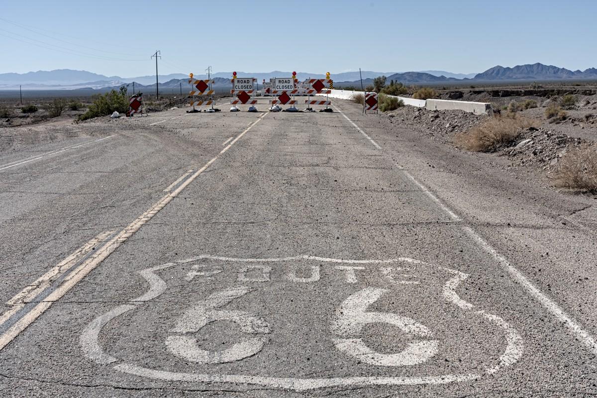 A color landscape photo of a highway going from the bottom of the frame to the top. The road is grey with a solid yellow line on the left and to the left of the solid line is a dashed line. Ahead there are barriers that say, "Road Closed." It's a desert landscape with a range of mountains low on the horizon. The sky is clear and blue.
