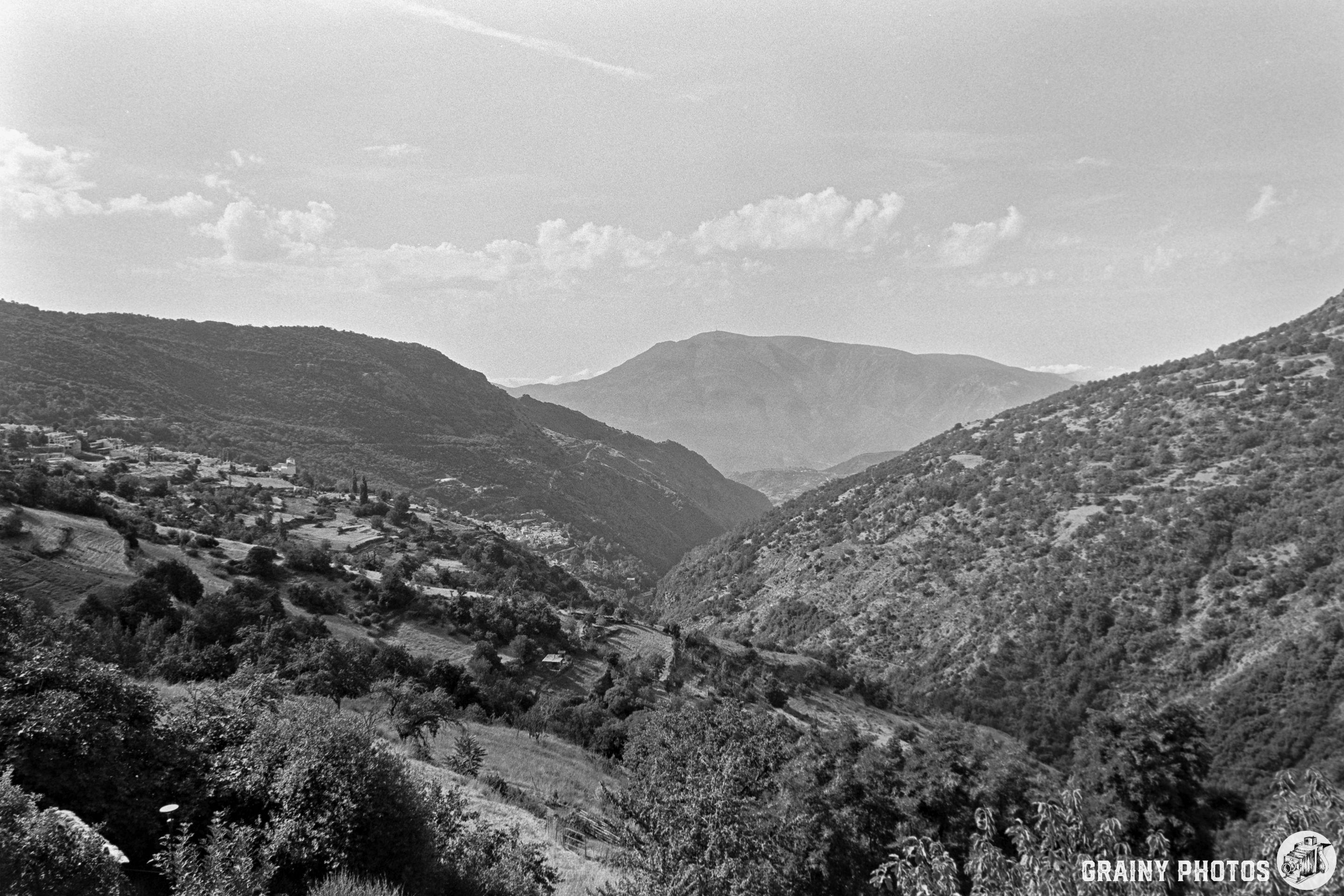 Black and white photo of the Poqueira Valley’s mountainous landscape with rolling hills and a distant peak under a partly cloudy sky. Vegetation and scattered trees cover the slopes.