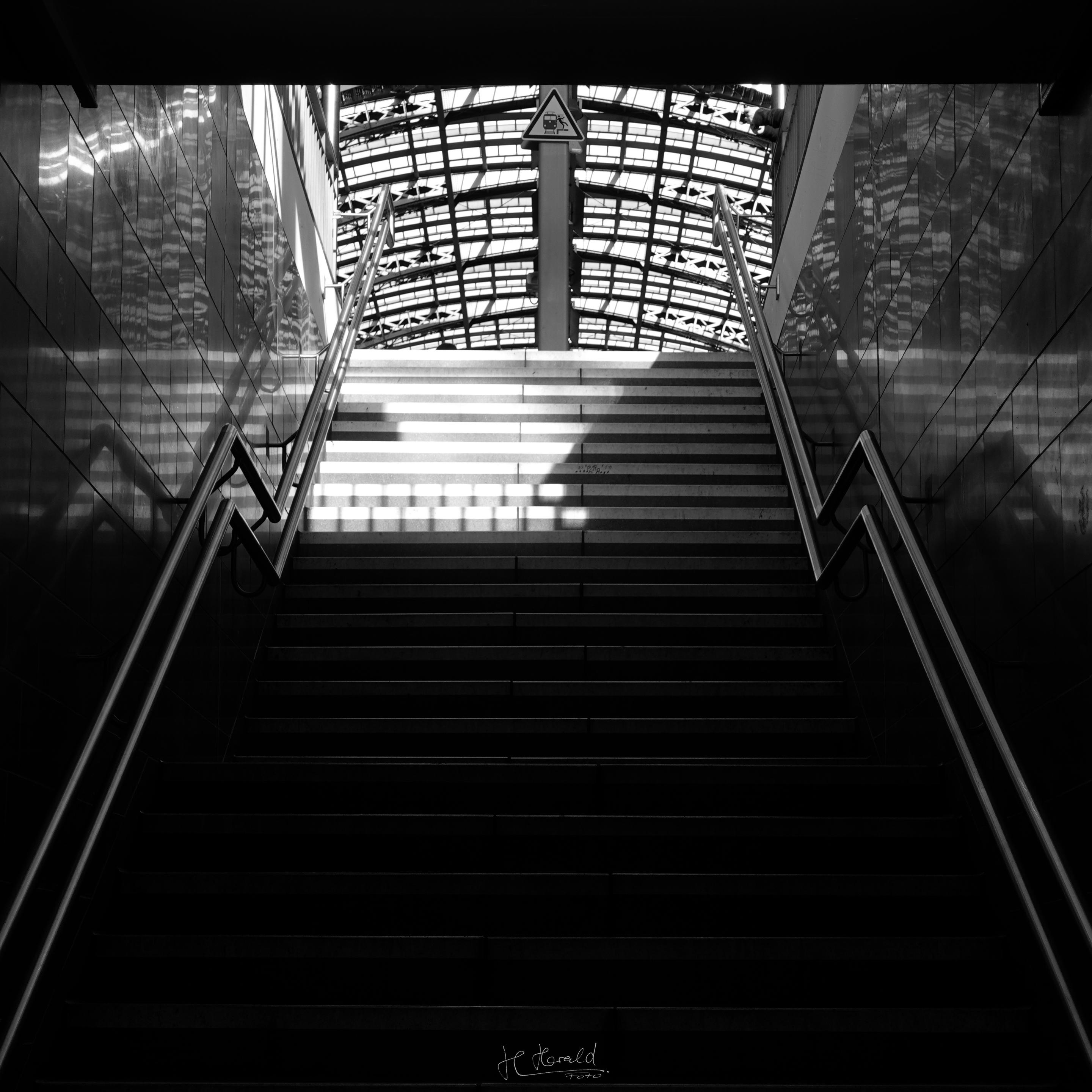 Square black-and-white image of a staircase leading into the hall of Frankfurt Central Station. The view goes up the staircase to the roof with its many skylights. The double metal railings on each side of the staircase have a bend at one landing. At the top centre of the image is a triangular warning sign with a train symbol. The sun shines from the right and illuminates the upper left staircase. Details of the roof are reflected in the clad walls of the staircase.