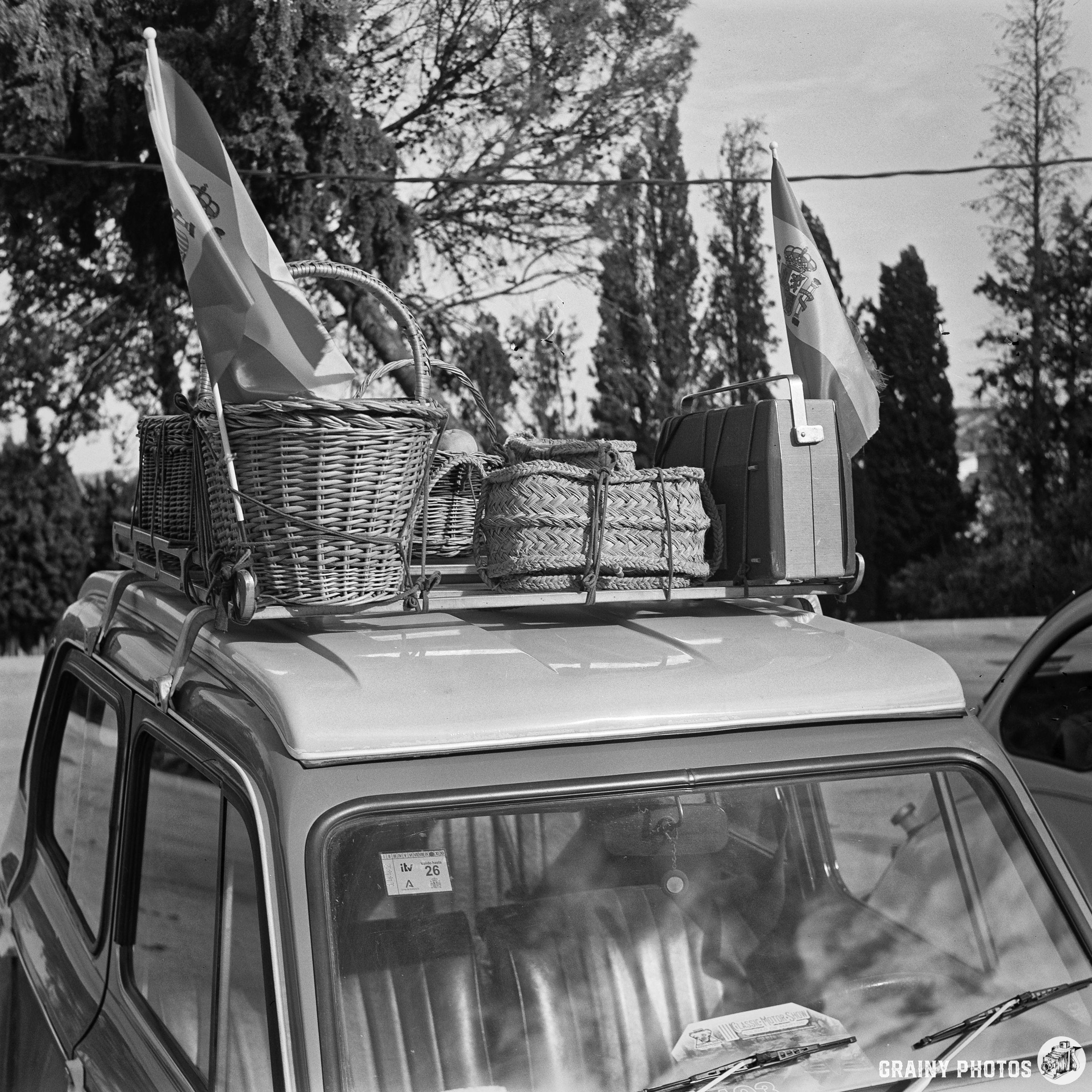 A vintage car with wicker baskets and decorative flags on its roof, set against a backdrop of trees in black and white. The scene conveys a nostalgic, leisurely atmosphere, possibly for a picnic or outdoor excursion.