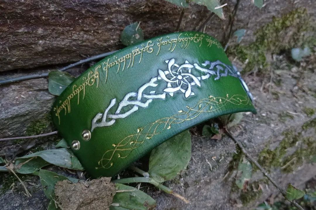 Partially sided pic of green leather cuff on a stone setup and some leaves, with various designs of The lord of the rings pieces: at the center, the Nenya design, the ring of Galadriel (a star shaped kind of flower, with intertwined branches at both sides) painted in metallic silver. At the upper border, the One Ring inscription, and an irregular design of vines and small leaves, both handcarved and painted in metallic golden