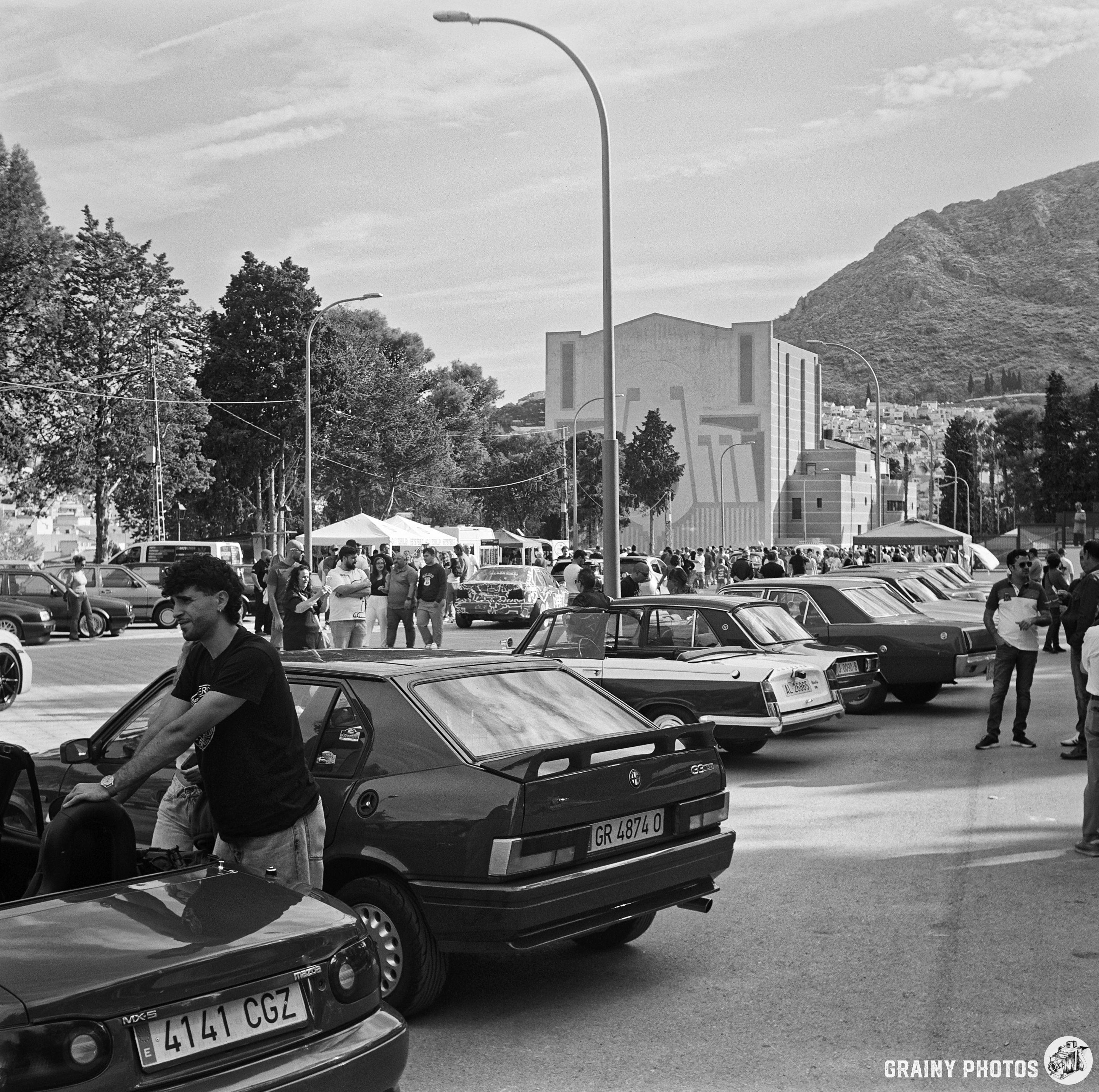 A bustling outdoor car meet featuring rows of vintage cars under a partly cloudy sky. People are interacting and enjoying the event, with mountains in the background and a modern building nearby.