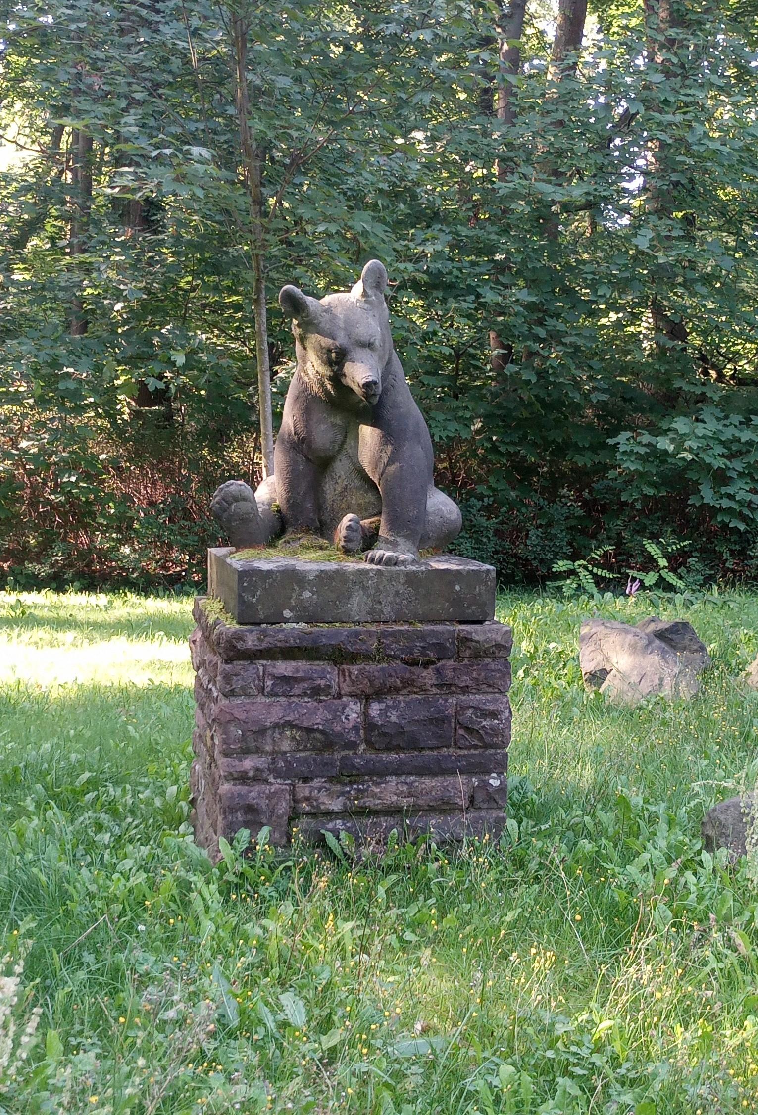 Statue of a bear sitting on its hind quarters on a metre high plinth of stones in green park surroundings. The bear has its head slightly tilted and an ear turned towards the viewer as if it's waiting for an answer.
