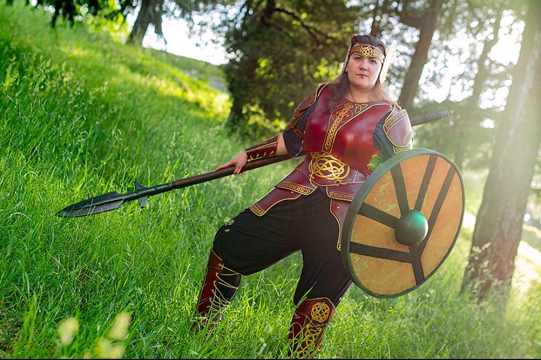 Horizontal pic of a woman on a green field wearing a leather full armor, an spear and a round viking shield. The armor is red with golden decorations of norse knots, a moulded semi-cuirass with a sword tooled on the center and a corset with two serpents intertwined, full golden tiara and bracers and greaves adjusted with golden cord