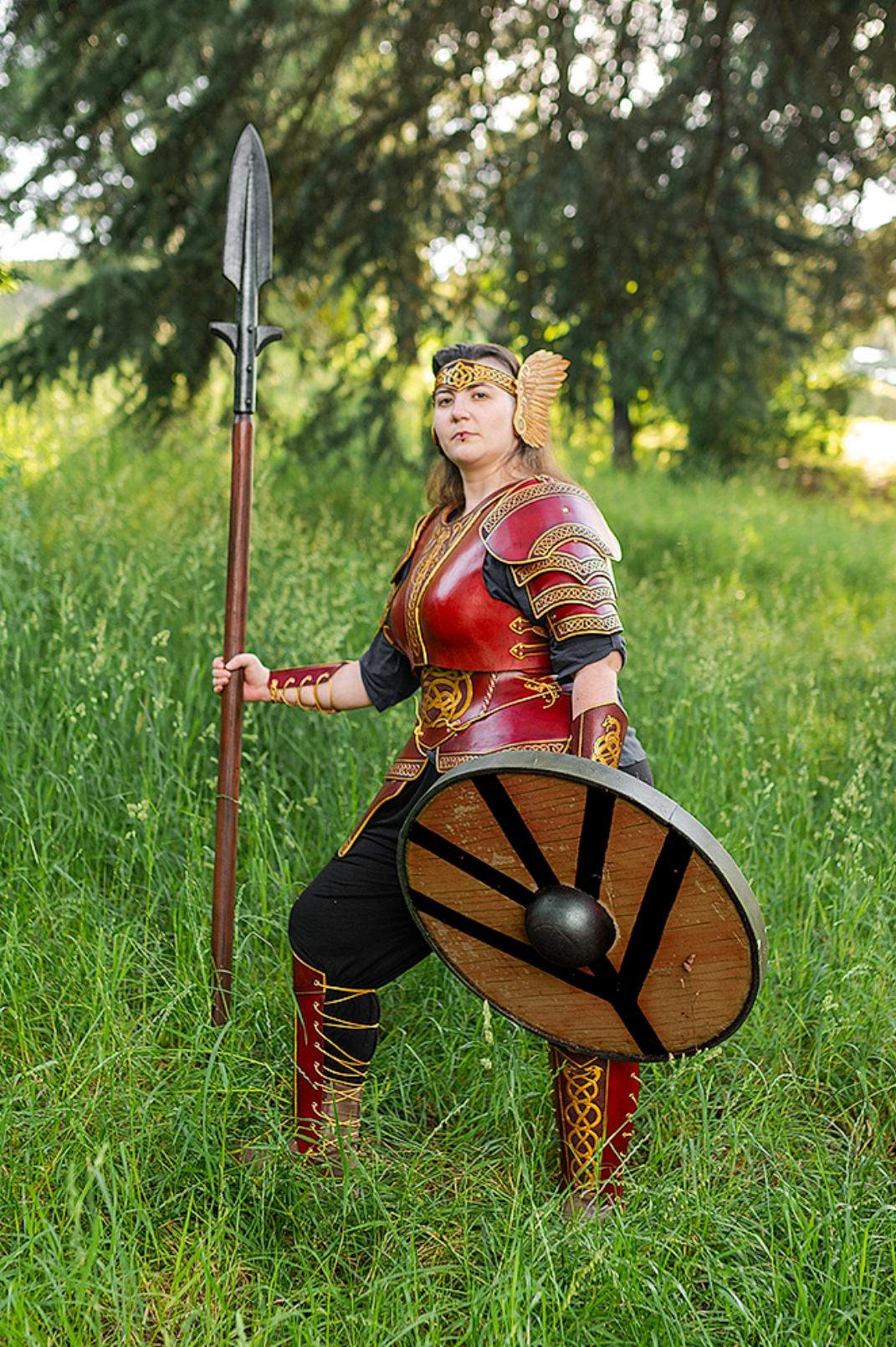 Another pic of the same woman as the first pic, on the same  green field, this time in a more relaxed posture, the spear resting, the shield facing to the ground, and the woman more side faced