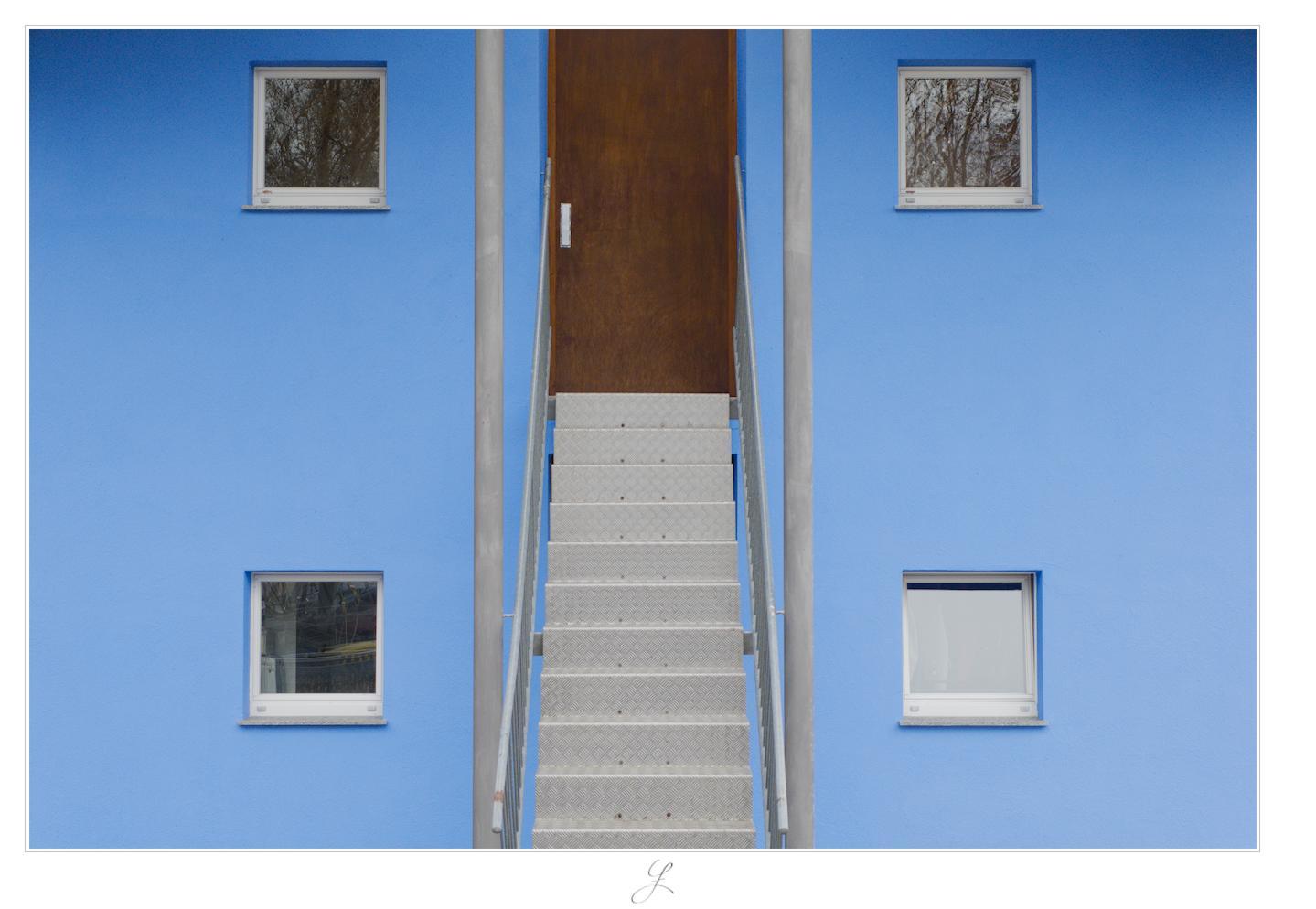The facade of a blue house, right upfront. It is quite symmetrical: in the center top, there is a silver-gray metal stair leading straight to a dark brown wooden door at the upper end. Left and right to the steps there are silver-gray metal railings. At the bottom of the stairs, there are two thin gray metal pillars at the end of the railings reaching from the ground straight up to the roof, one to the left and the other to the right. Left and right of the pillars, four small symmetrically ordered squared windows can be seen, each of it exactly marking the four corners of a larger square reaching from the ground floor to the first floor (perhaps not exactly a square, but it seem as if it could be a square).
AI disclaimer: Using my work, its meta data, written or derived description to create media with or train AI based systems is prohibited.