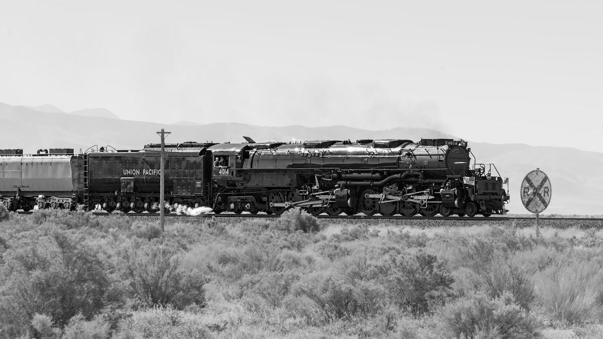 A black and white landscape photo of a very large black steam engine in a desert landscape. The engine is heading toward the right side of the frame. It is very long, with two sets of drive wheels. The tender car is behind the engine and a small section of a passenger car can be seen behind the tender. Sagebrush is in the foreground, and dimly, through the haze, a mountain range is seen in the distance. A round, railroad crossing sign is seen just ahead of the engine on the right. It is filled with bullet holes.