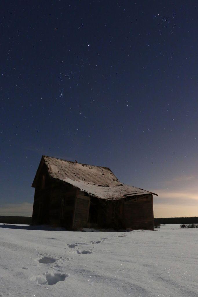 Night-time photo of a dilapidated country home in the winter. Snow stretches in the foreground with footprints leading to the house. Above the house is a clear night sky, boasting the constellation of Orion against a smattering of other stars. There is some mild light pollution on the horizon--occupational hazard of photographing near a large city.
