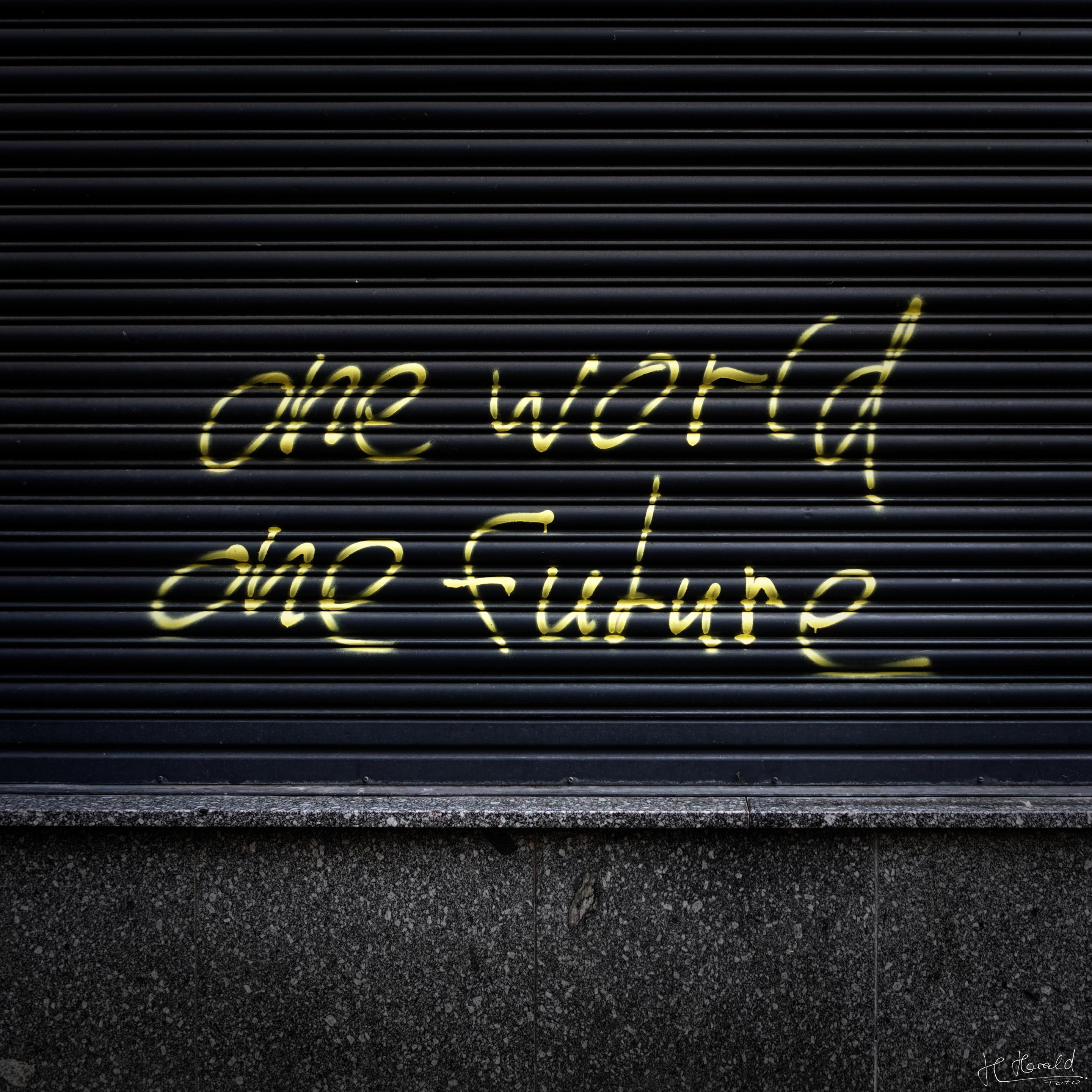 Yellow lettering on a black roller shutter covering the upper two-thirds of the image. The lower third shows a stone façade. The text reads ‘one world, one future’. 