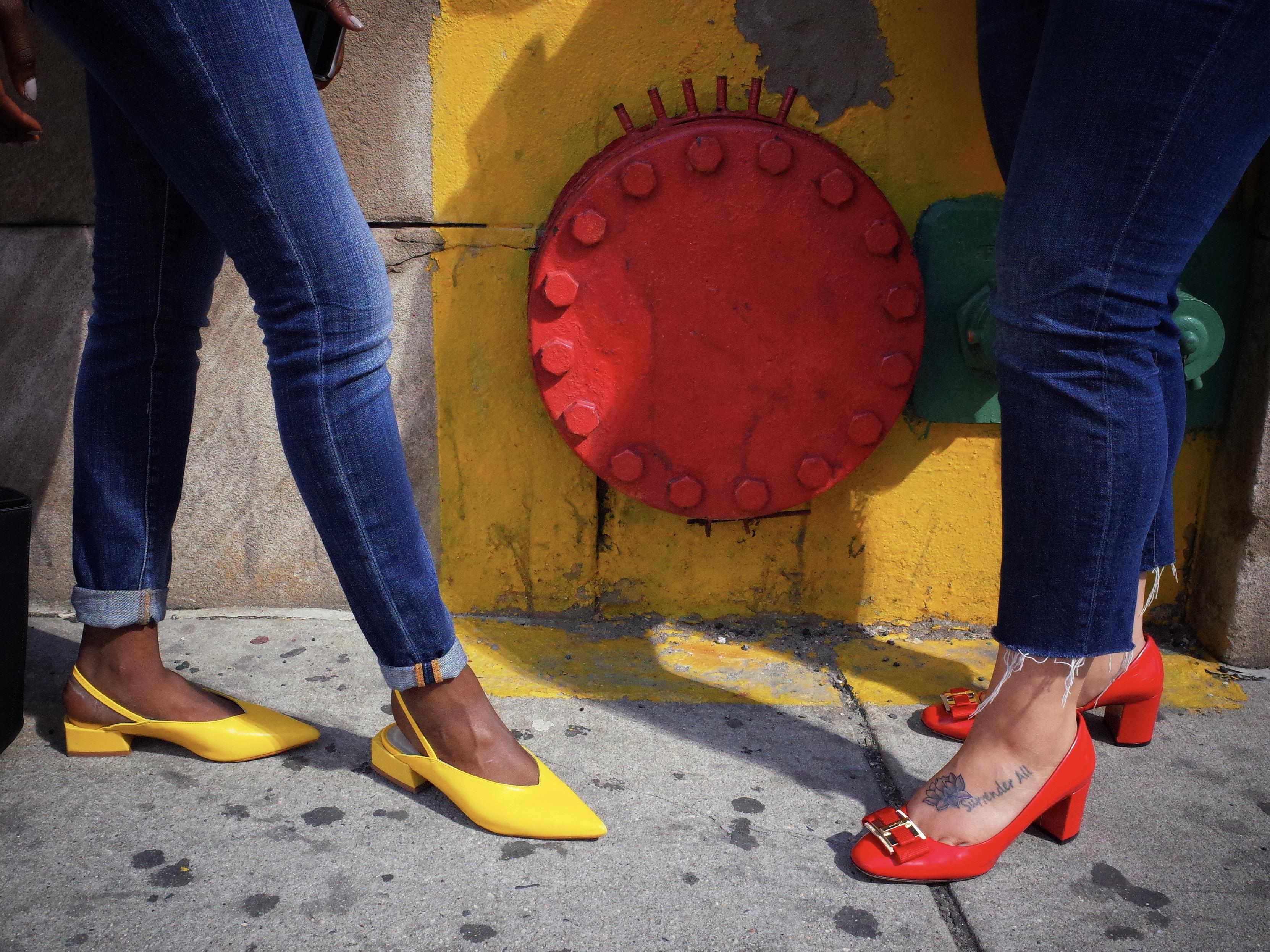 Two young women, one wearing yellow shoes and the other red shoes, stand in front of a red metal street drain which is surrounded by yellow paint. Both women are wearing blue denims.
