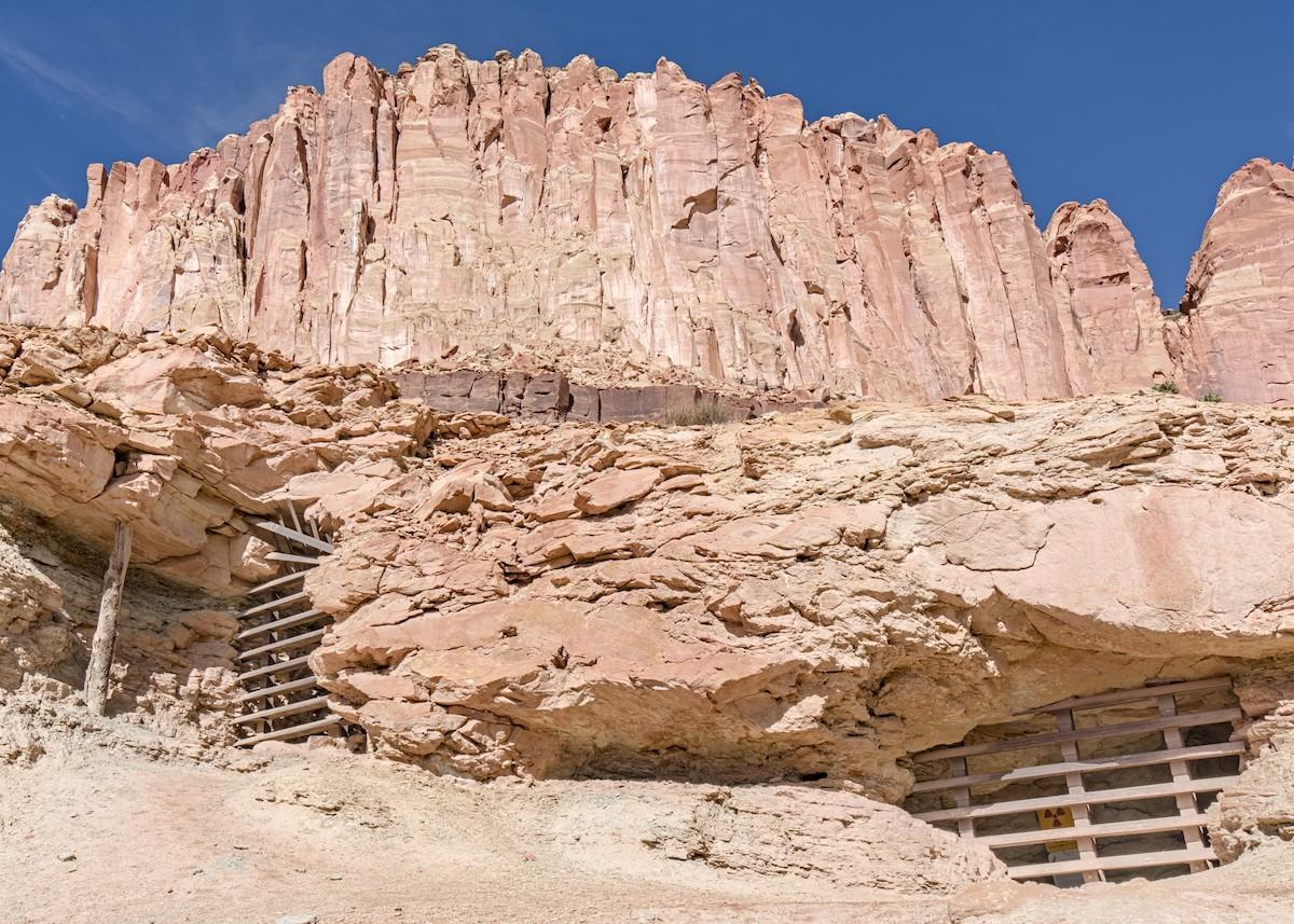 A color landscape photo of a rugged vertical light-red sandstone cliff face. In the foreground is a short cliff maybe ten or twelve feet high. Underneath a shelf like overhang are mine openings on the right and on the left that have been closed with iron bars to keep people out. Looking closely, a yellow sign with a red three leaf flower like design sign can be seen inside the mine opening. It is a sign that warns of dangerous radiation levels inside of the mine. Above and behind the shelf like overhang is a tall, hundred foot-plus sheer cliff. The sky is clear and bright blue.