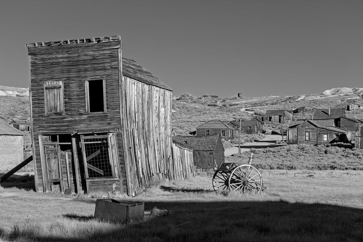 A black and white landscape photo of a ghost town. On the left is a very weathered two story wood plank building. It has two large windows on either side on the upper floor. There's a large window on the right on the ground floor with a door on the left. A large rusty metal box sits in front of the building. On the right side is a two wheel horse cart. The building is very weathered and the wood is wrapped and it leans severely to the left. In the background on the right is a dirt road going up a hill with numerous long abandoned wood buildings lining the road. The sky is clear and a dark gray as a result of processing. A shadow  from a building behind the camera is seen in the lower right.