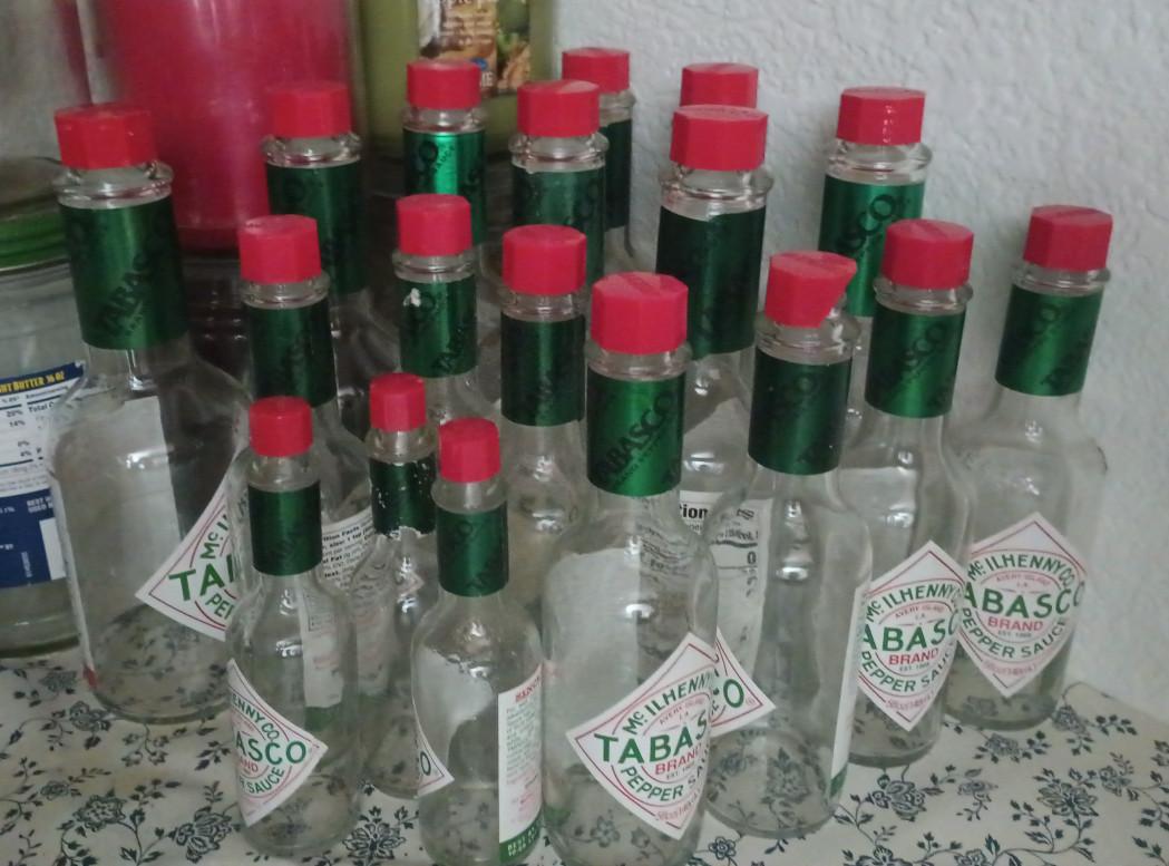 A color landscape photo of large, medium, and small bottles of Tabasco brand red pepper sauce. There's about twenty bottle total in the image. They are a narrow cylindrical shape with red plastic screw on lids.