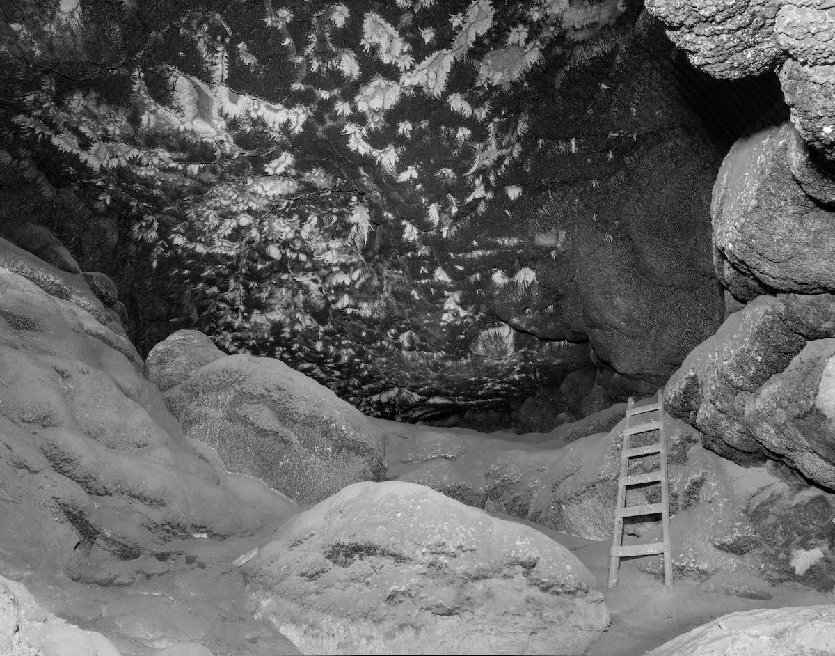 A black and white landscape photo of the inside of a cave. There are large boulders in the foreground and on the right. A six steps wooden ladder is on the right leading form the floor of the cave to a shelf in the back. The roof of the cave is covered in soot from decades of fires built by prehistoric Native people. The soot has light colored patterns in it that look like fern leaves possibly due to the growth of a fungus or mold. 
