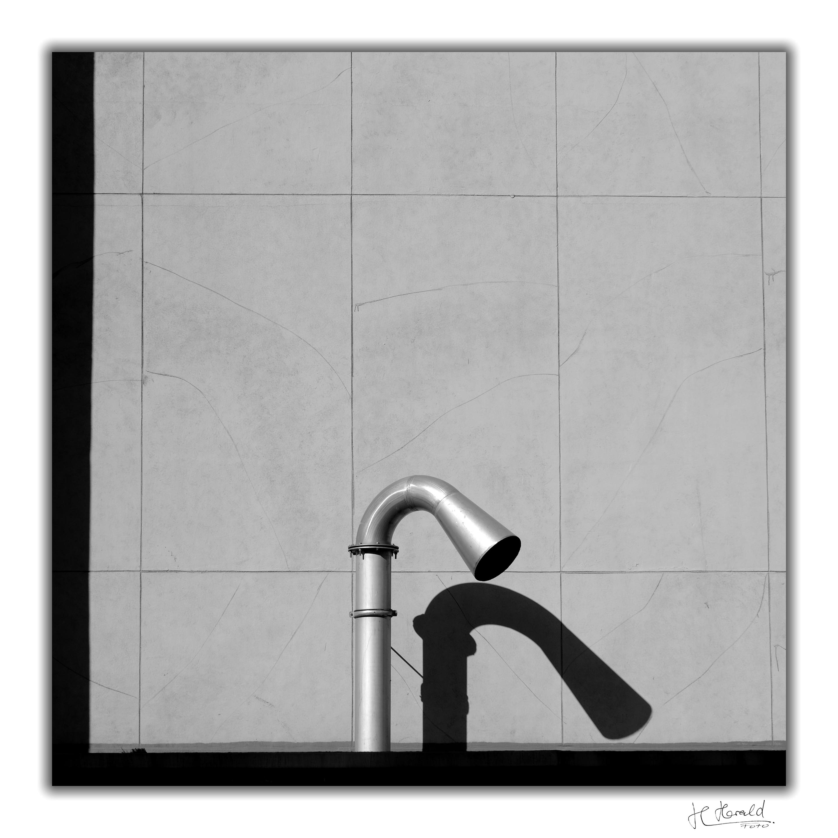 Square black-and-white image with a white frame of a bent pipe. The wide metal pipe enters the frame from below and stands in front of a grey wall made of large rectangular stone slabs. It curves diagonally downwards at the top and opens slightly to form a sort of funnel. A distinct shadow of it is visible on the wall. On the left and at the bottom of the frame is a broad, straight black shadow, like half a frame.