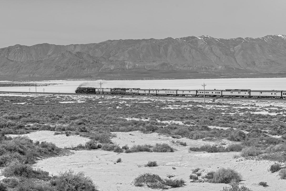 A black and white landscape photo of a large steam engine with about ten cars traveling across the frame from right to left. A bit of black smoke is coming out of the smoke stack. In the for ground is a light colored landscape dotted with clumps of desert shrubs. Behind the train is a stark white dry lake playa. Beyond the playa is a range of mountains with a tiny bit of snow on top.