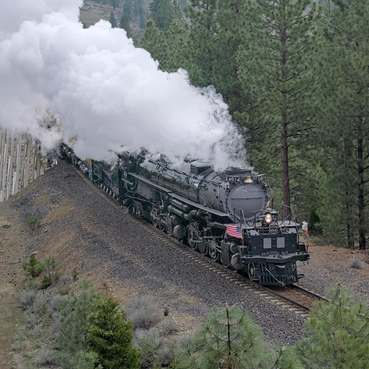 A color photo of a very large steam engine moving through a forest toward the lower right of the frame. An American flag flutters on the engine's right side. Very large billows of steam boil up and toward the upper left of the frame.