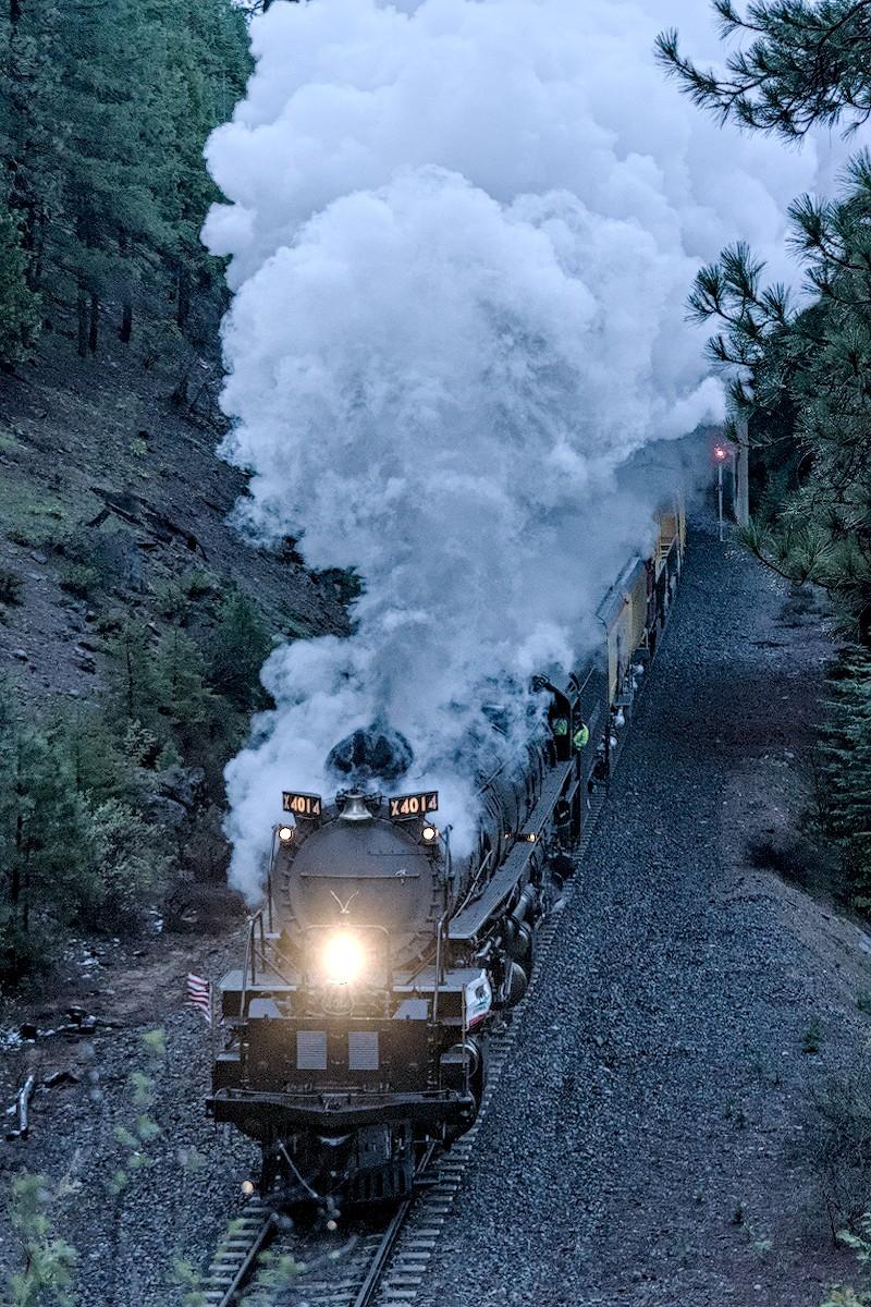 A color portrait photo of a large steam engine under full load chugging up canyon in the late afternoon. The view is from above as the train moves from the upper right to the lower left. The headlight shines brightly and at the top of the round boiler at the front of the engine are two small signs on either side of the central bell that say, "X4014." Gobs of steam and smoke boil up from the front smoke stack and fill the view to the top of the frame.
