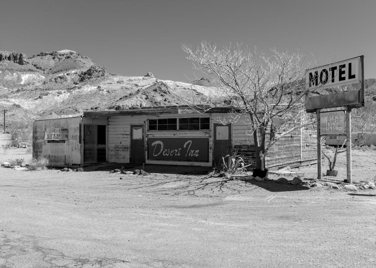 A black and white landscape photo of an old abandoned motel office. It's a pretty much rectangular wood-planked building that looks a lot like an old gas station, which it may have well been at one time. On the front, in cursive lettering, it says, "Desert Inn." We see the right corner of the building so the front is on the left and a windowless right side. A small dead tree stands at the corner. On the far right is a "Motel" sign that is a little above roof height. There's a flat gravel lot in front and in the distance, behind the building, is a barren desert mountain on the left.