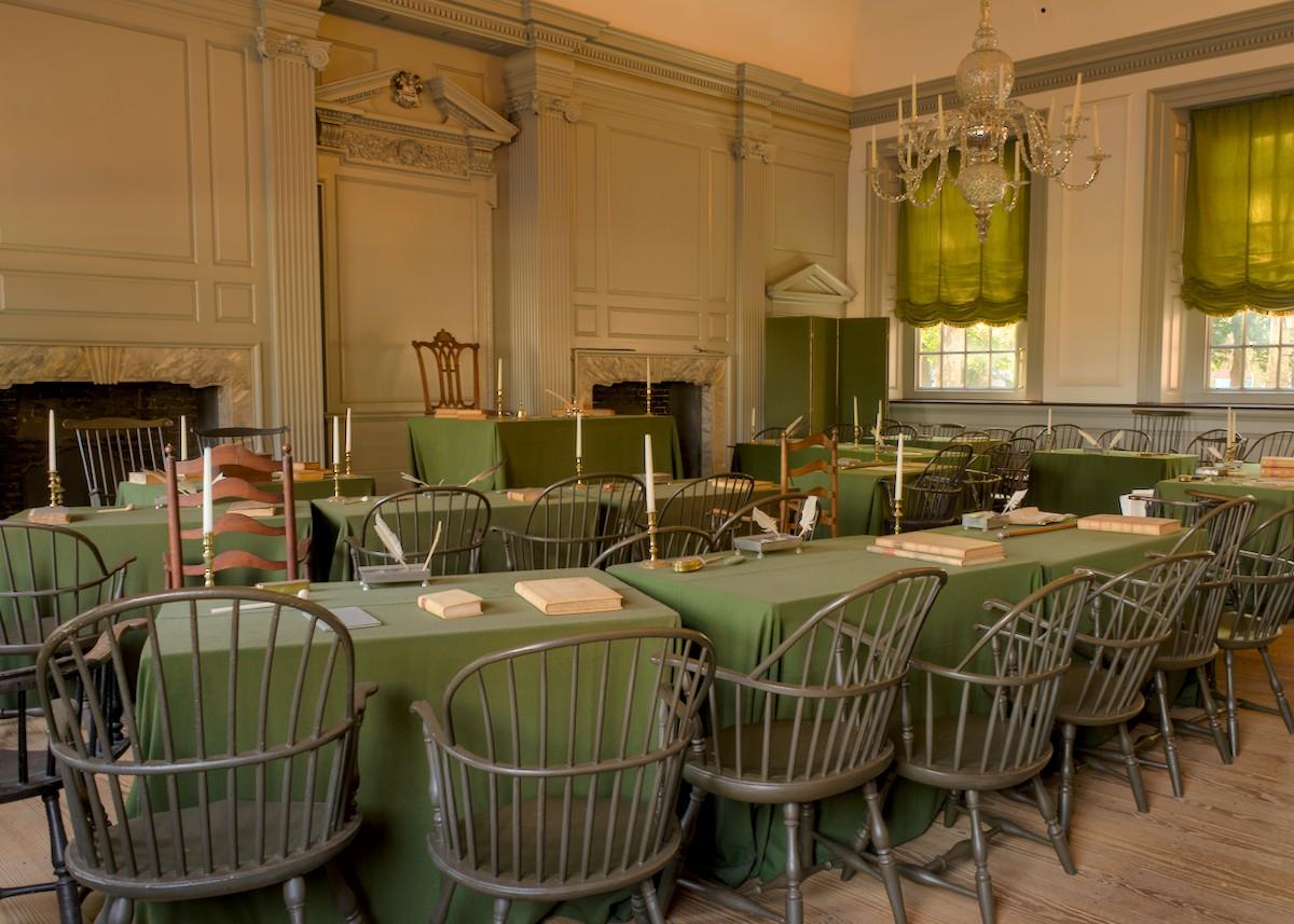 A color landscape photo showing the room inside Independence Hall in Philadelphia where where both the Declaration of Independence and the Constitution of the United States were debated and adopted by the Founding Fathers of the United States. The room features Georgian architecture with faux columns and carved masks and scrolls. Two rows of tables with muted green felt table cloths, books, candles, and quill pens, face a single table on a raised area in the back of the room. George Washington's "Rising Sun Armchair" is facing the room behind a small table on the riser. Dr. Benjamin Franklin wrote: "I have often looked at that picture behind the president without being able to tell whether it was rising or setting. But now at length I have the happiness to know that it is a rising and not a setting sun." Let's ensure that sun raises again--VOTE!