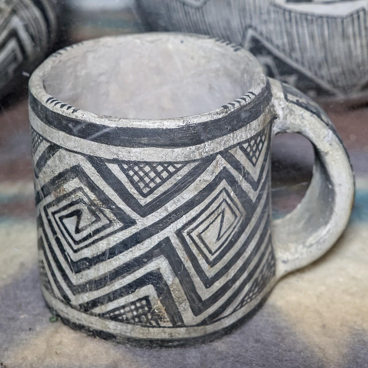 A square color photo of a black on white pottery mug. It looks exactly like a well made pottery mug you might find at any modern day pottery. The black line decoration is made up of connecting zig-zag diamond shapes. There is a bit of similarly decorated pottery out of focus behind the mug in the foreground.