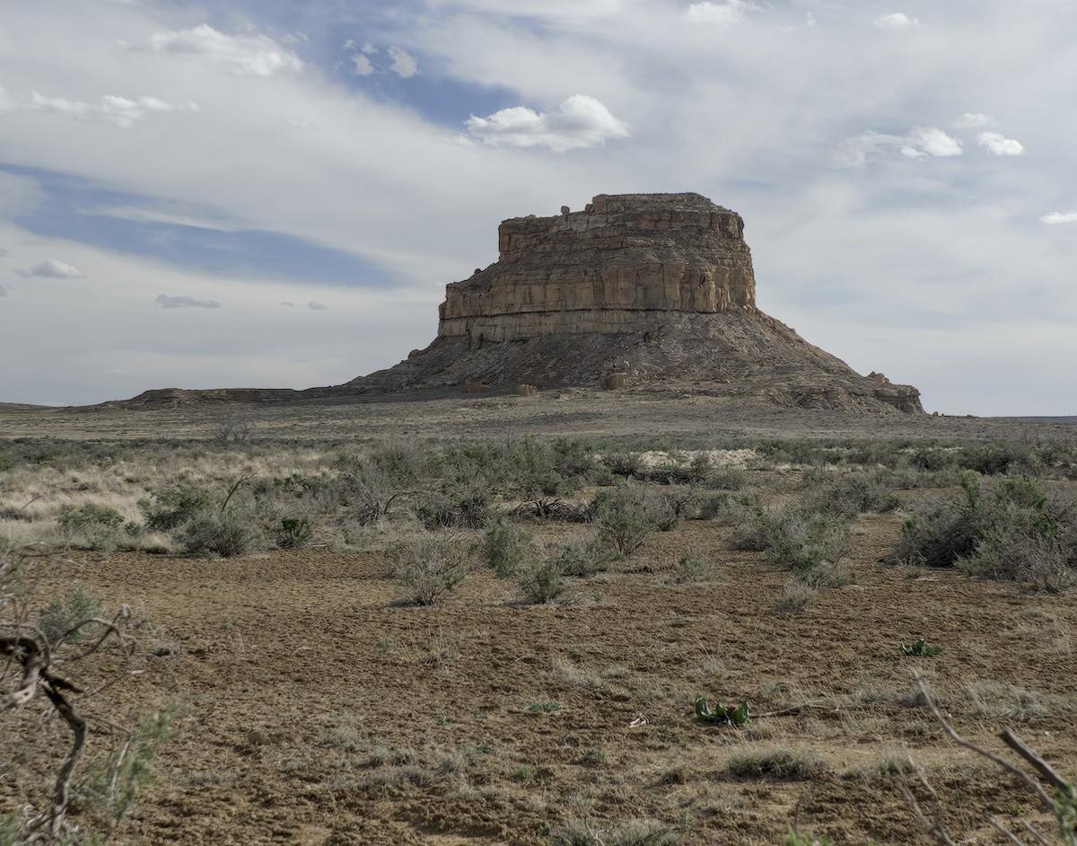 A color landscape photo of a tall solitary round butte on a high desert plain of small shrubs. The sky is mostly cloudy with small patches of blue sky.