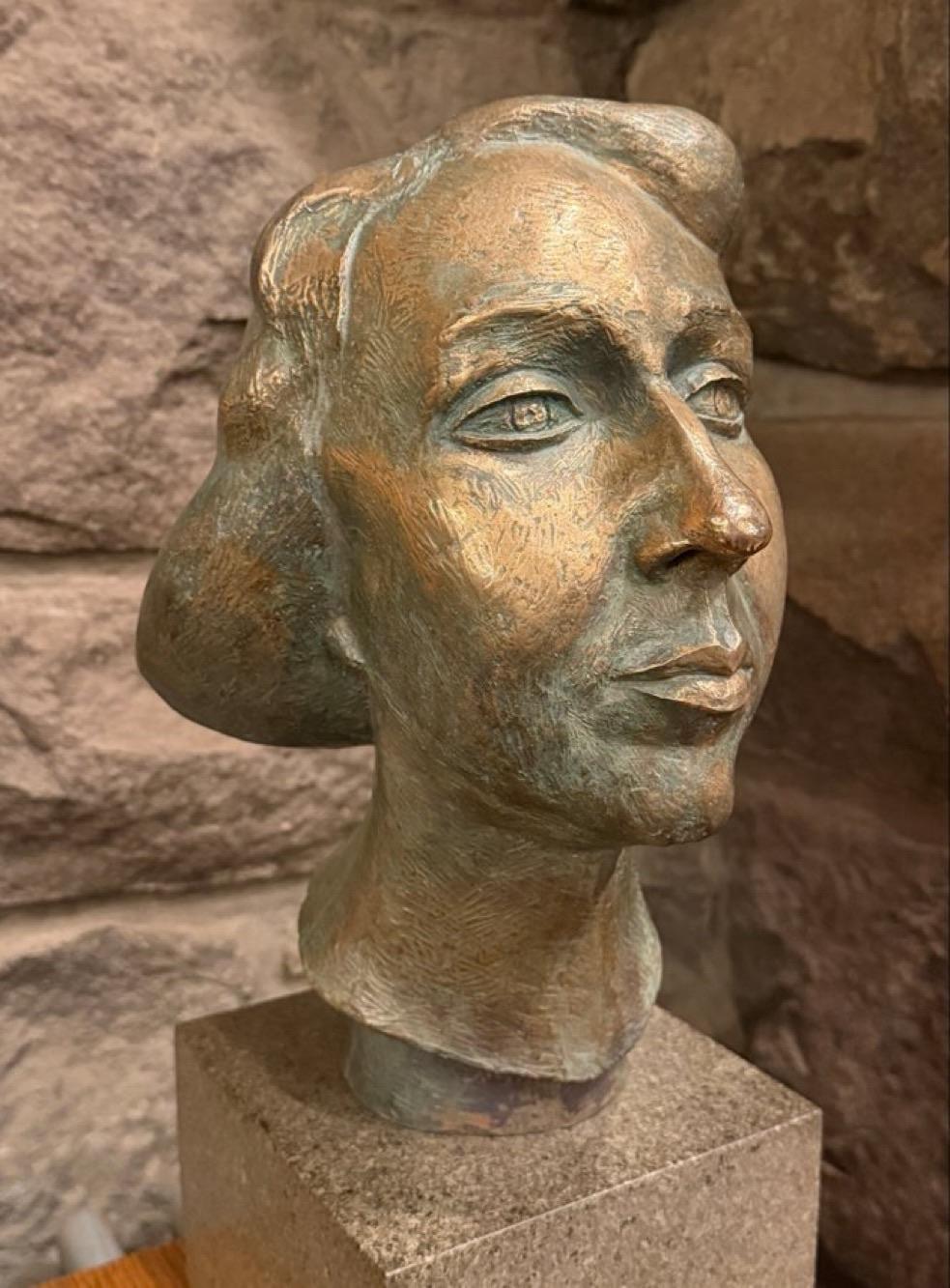 A bronze sculpture of a woman's head, featuring detailed facial features and a smooth finish. The sculpture is mounted on a square, textured base and is set against a stone wall background.