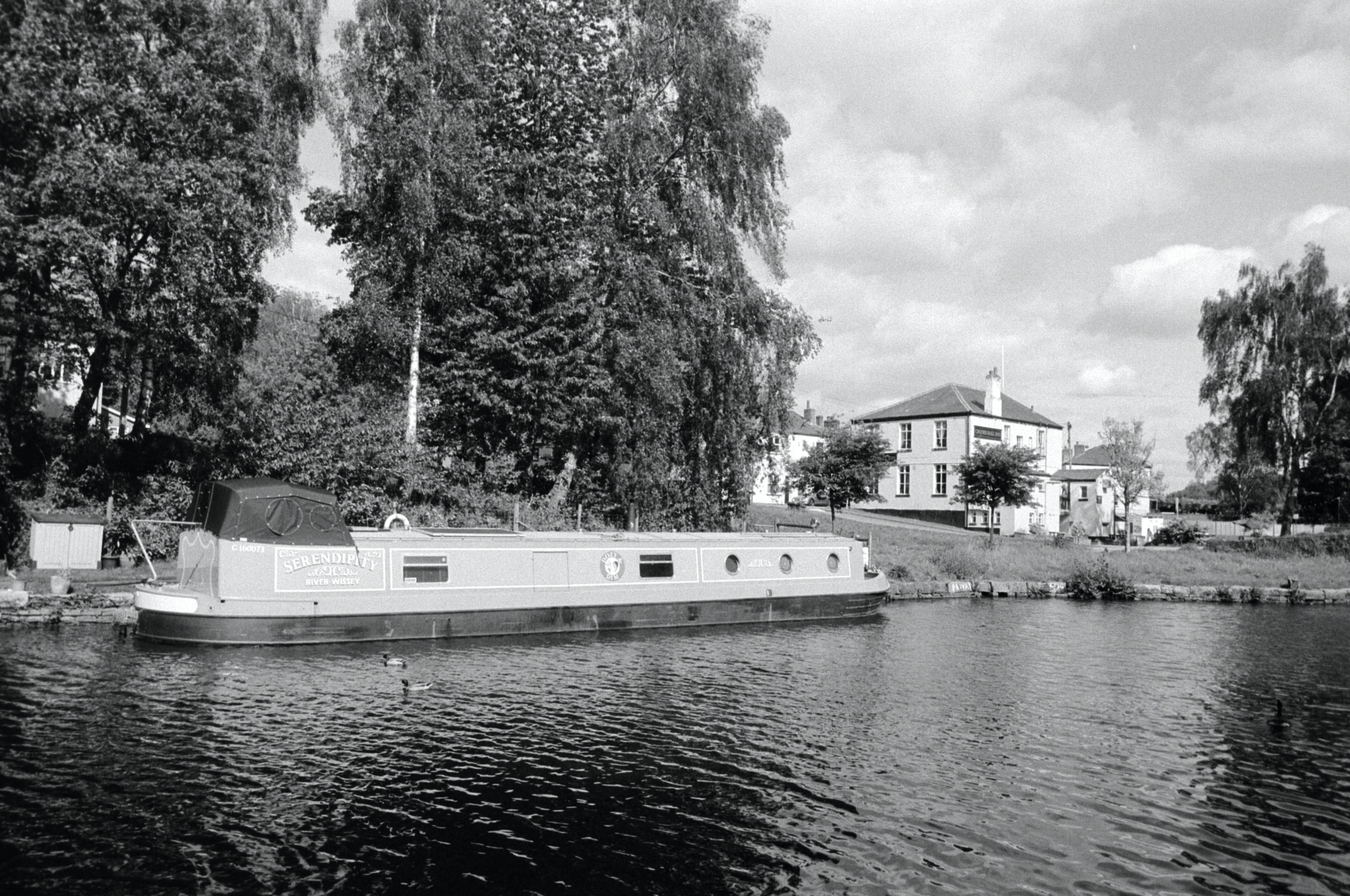 A black and white film photo of a narrowboat moored on the Leeds and Liverpool Canal with the Crooke Hall Inn in the background to the right. It's a lovely place to live.