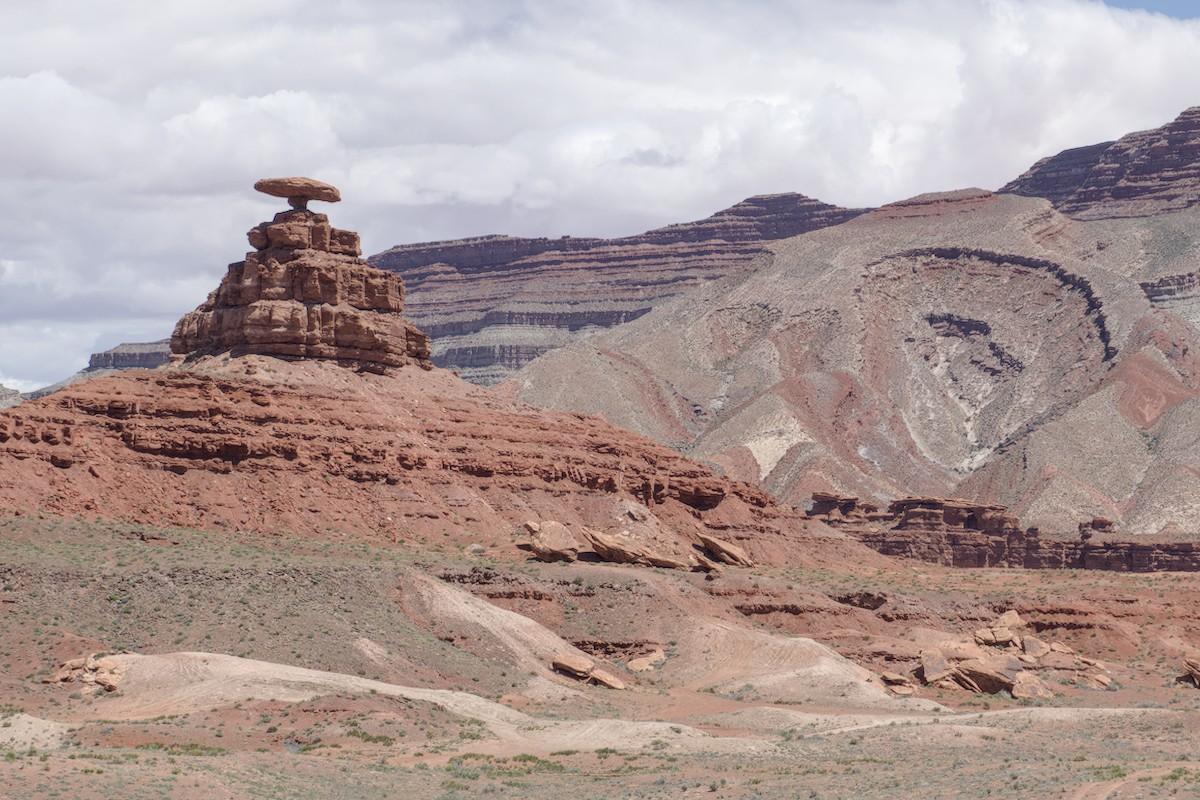 A color landscape photo of red and gray sandstone formations. On the left is a conical shaped hill eroded into three steps near the top with a large disk shaped rock balanced on the fourth tinyist step at the top. It resembles a Mexican sombrero. On the right is a large hill with zig-zag layers of rock at the base and a large crater like feature on the side. In the background is a mountain showing straight horizontal layers of rock. The sky is overcast.