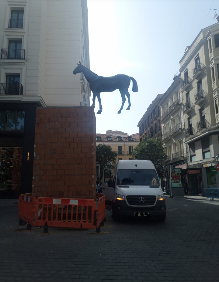 My photo of a street with a bronze horse statue high up in the air with a minimal connection to a bricked up pedestal. The surroundings are normal street live continuing as if not noticing or caring.
