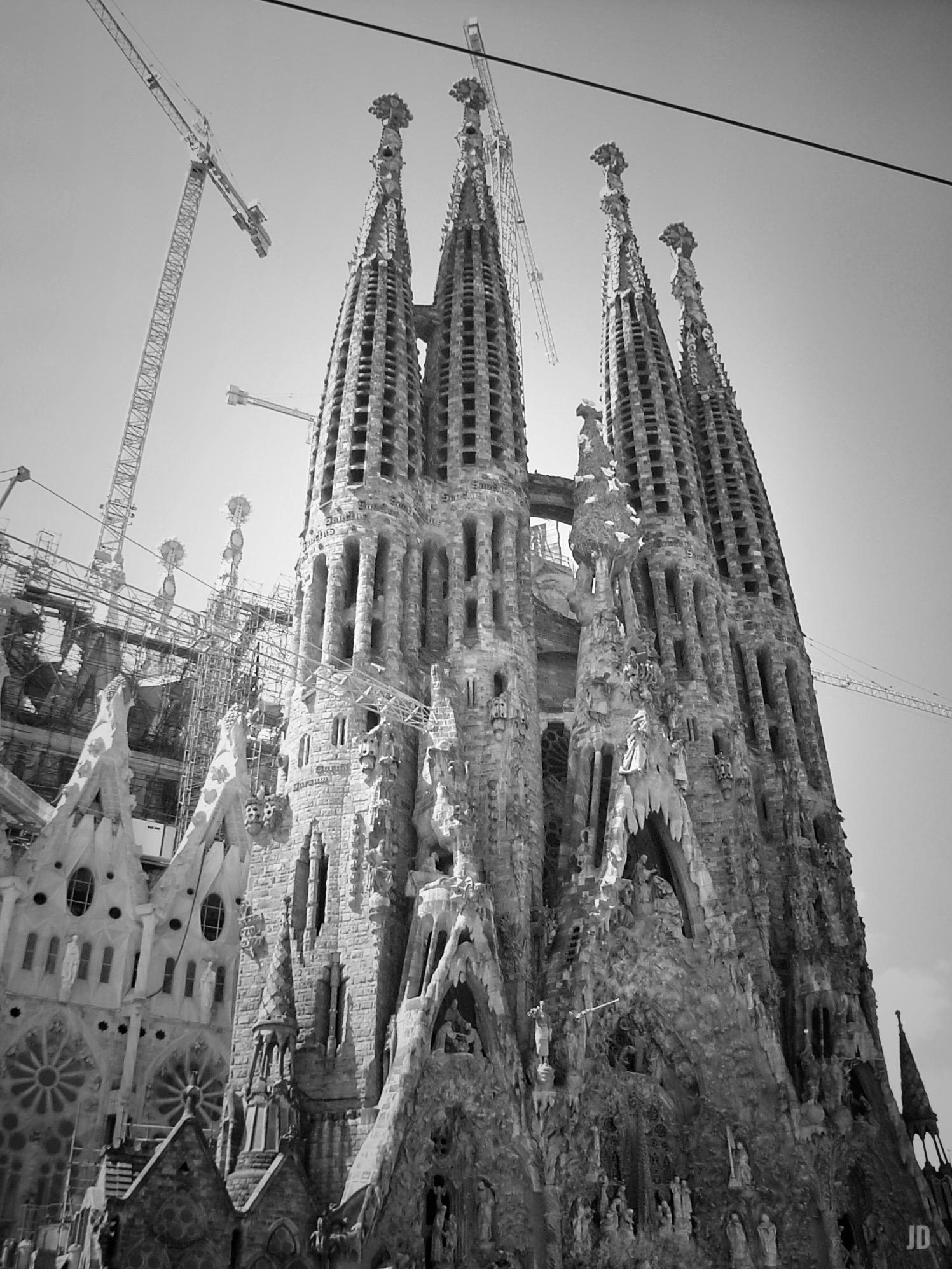 La imagen muestra la fachada principal de la Sagrada Familia, la famosa basílica en Barcelona diseñada por Antoni Gaudí. Se aprecian varias de sus altas torres con formas orgánicas y detalladas esculturas, mientras varias grúas indican que el edificio sigue en construcción. La foto está en blanco y negro, resaltando los contrastes de luces y sombras en la piedra.