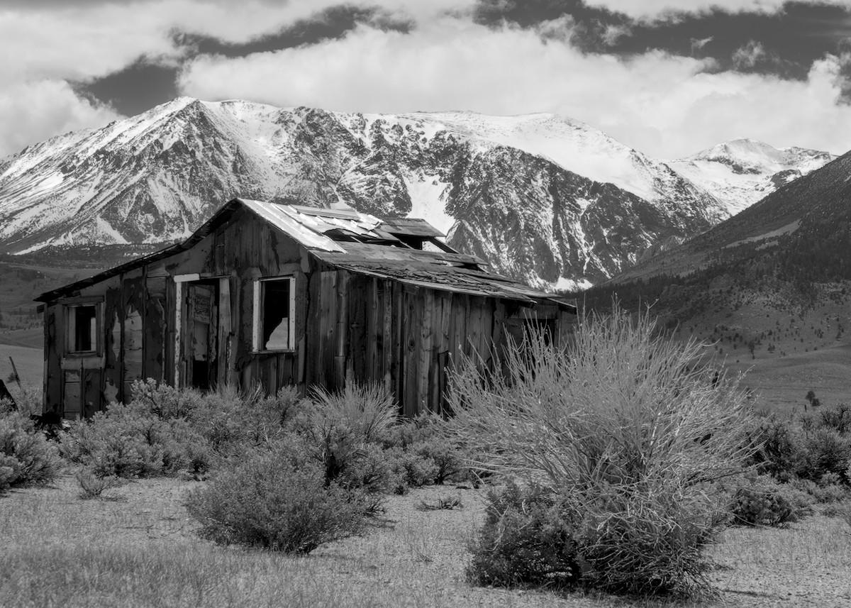 A black and white landscape photo of an abandoned cabin in a sagebrush plain with tall snowy mountains in the background. To the left, the cabin is in poor shape and without windows or a door. There's a hole in the roof as well. The sky above the mountains is mostly cloudy and the tops of the mountains are obscured by the clouds.