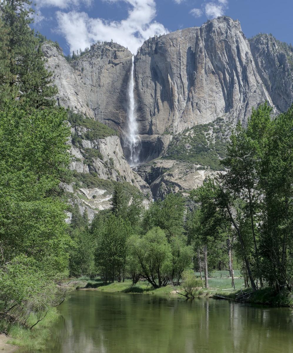 A color portrait photo of a tall waterfall in the distance pouring over a solid gray granite rock face. There's a "V" shape in the rock face that the water fall is pouring from and down. It disappears behind more rock formations about two thirds of the way down. In the fore- and middle-grounds is a very still looking green river and trees of various sizes and types. A bit of a meadow opens up on the right middle-ground. The sky is bright blue with puffy white clouds.