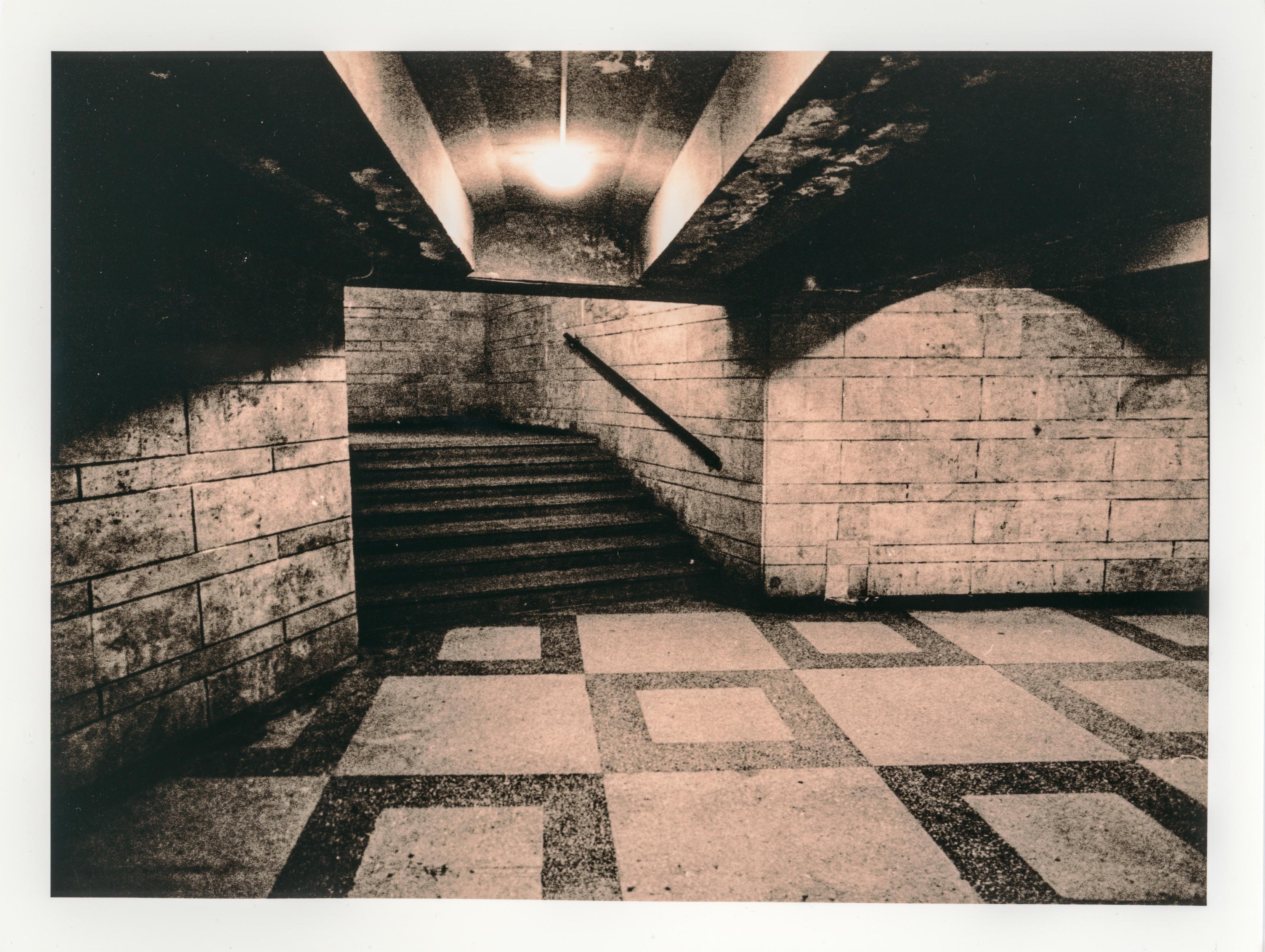 Lith print of an empty underground passage in central Lviv. A staircase disappears into shadow beneath a single glowing light, while textured stone walls and patterned floor tiles create a stark, moody composition.