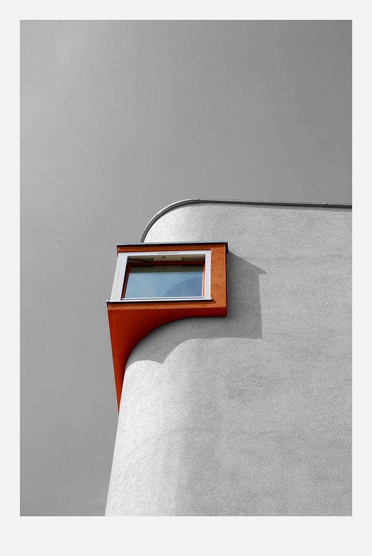 Eine abgrundete Hausecke nach oben gegen den Himmel fotografiert. Aus der Rundung steht fast oben eine orangerote Ecke hervor, in der ein Fenster ist.
//
A rounded corner of a house, photographed looking upwards towards the sky. An orange-red corner, containing a window, protrudes from the curve near the top.