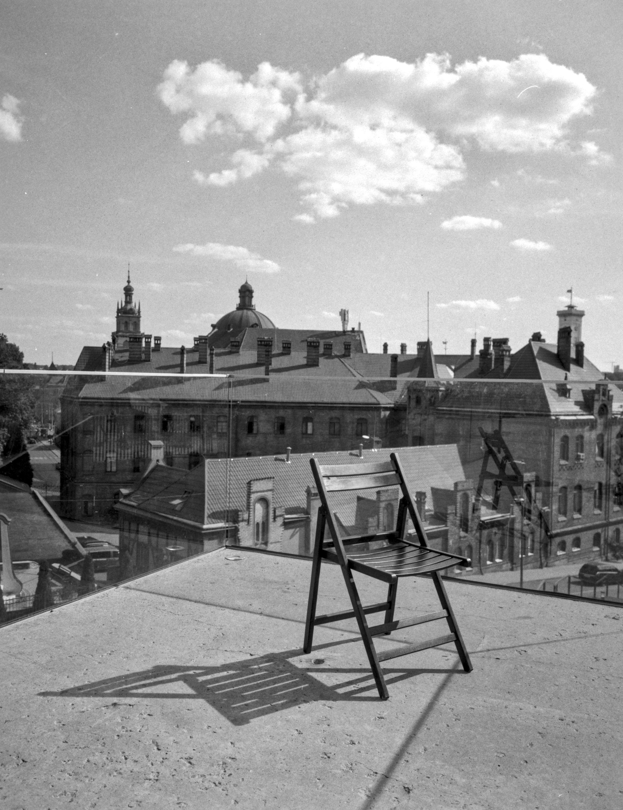 A black-and-white view from a terrace in Lviv, where a single folding chair faces the historic skyline. Church towers and old rooftops are seen through a clear glass railing beneath scattered summer clouds, while the chair casts a sharp shadow across the sunlit concrete.