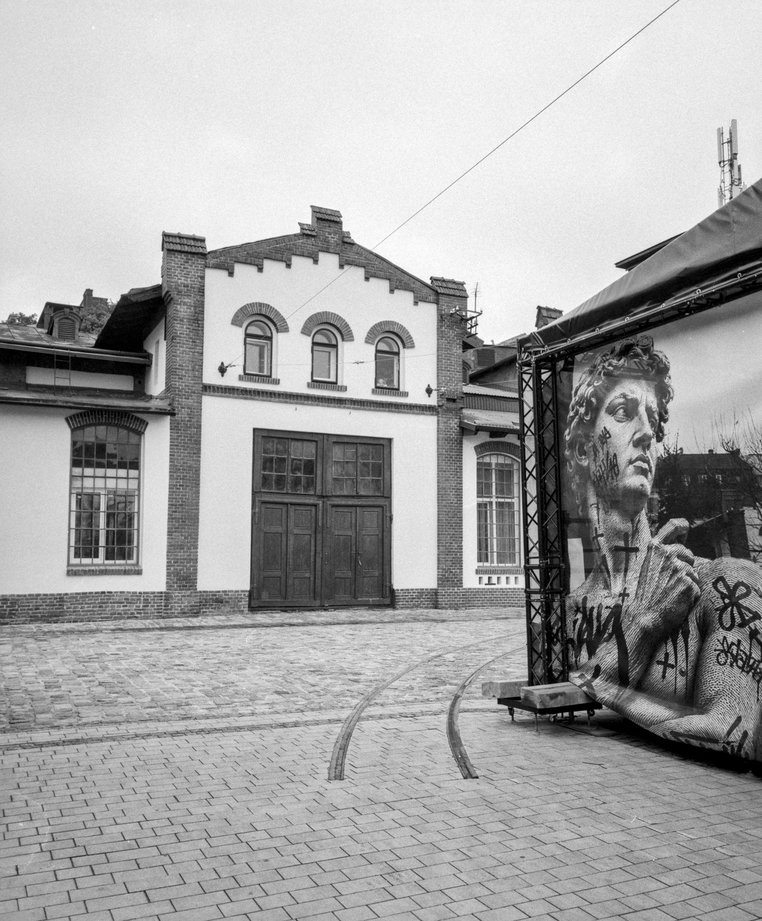 Black-and-white photograph of a historic brick industrial building in Lviv with large arched windows and a central double door. Curved narrow-gauge rails embedded in the paved courtyard lead toward the building. On the right, a large mural reproducing Michelangelo's David covered with graffiti-style markings stands on a temporary exhibition wall, contrasting with the restored architecture