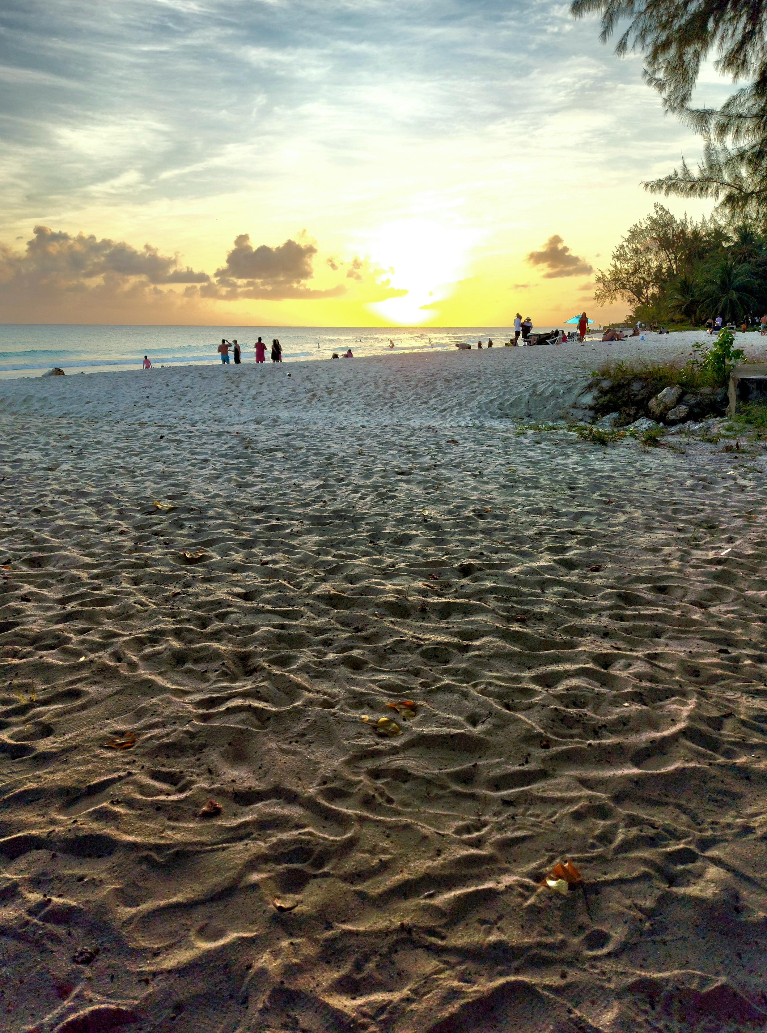 Sunset on a beach in Barbados. Small clouds on the horizon and, far off on the beach by the water, the silhouettes of several people. To the right, local vegetation. The majority of the photograph is taken up by the beach sand, which, backlit by the setting sun, creates a beautiful visual texture.