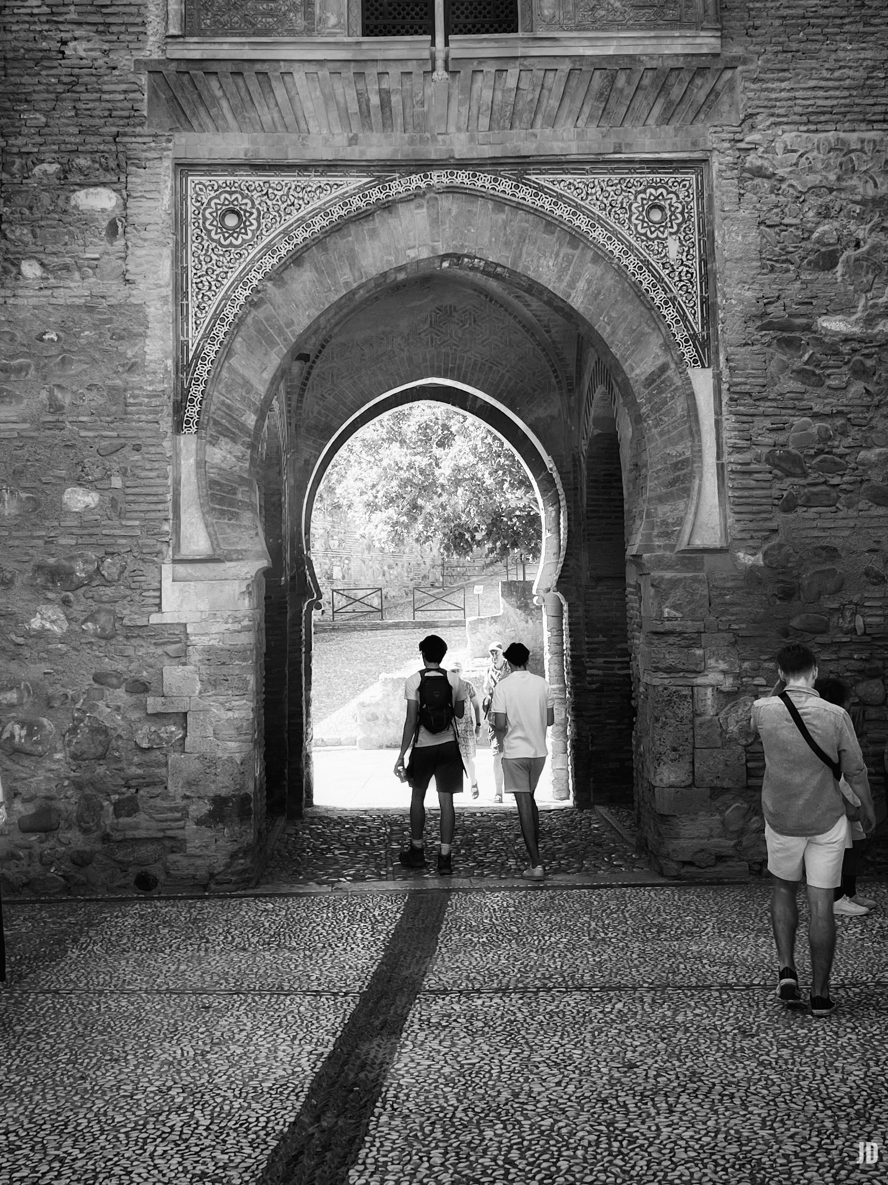 La imagen muestra una fotografía en blanco y negro de un antiguo arco de piedra con detalles ornamentales en la parte superior, posiblemente de estilo mudéjar o islámico. Por el arco, que sirve como pasaje, caminan tres personas de espaldas, mientras la luz exterior ilumina el fondo, donde se distingue un árbol y parte de un camino empedrado.