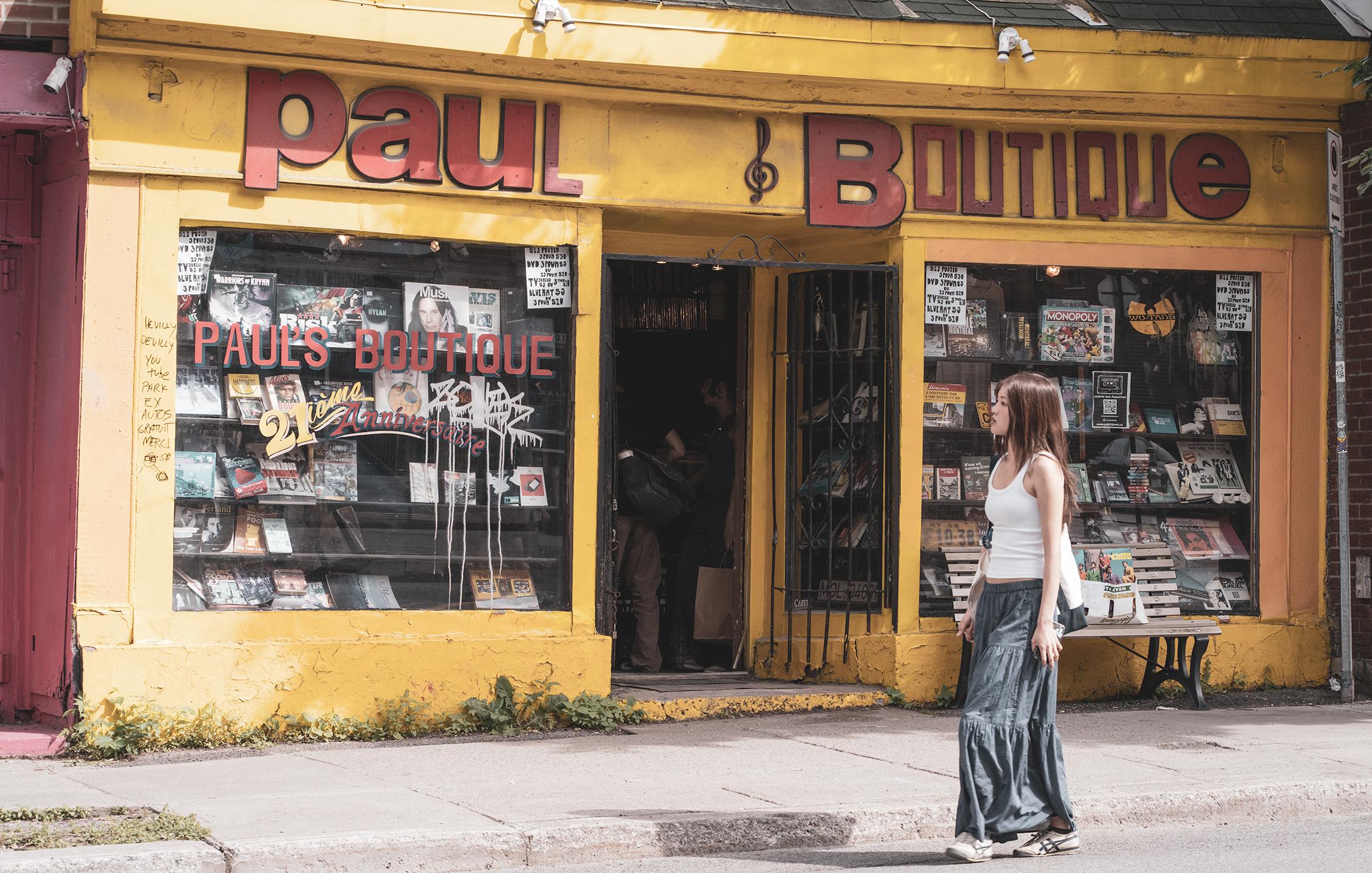 An open metal gate-style door invites passersby inside a yellow painted store front featuring bold, irregular red letters - Paul Boutique - and a treble clef. Two large windows display samples of the wears inside: vinyl, cds, games, etc. In between the windows, an alcove and a dark entry in which customers can be made out. In front of the store, a sidewalk from which a few weeds spring here and there, a park bench with a few records on sale, and a passerby: a young woman with long dark hair, a white tank, long blue-grey skirt, and sneakers.