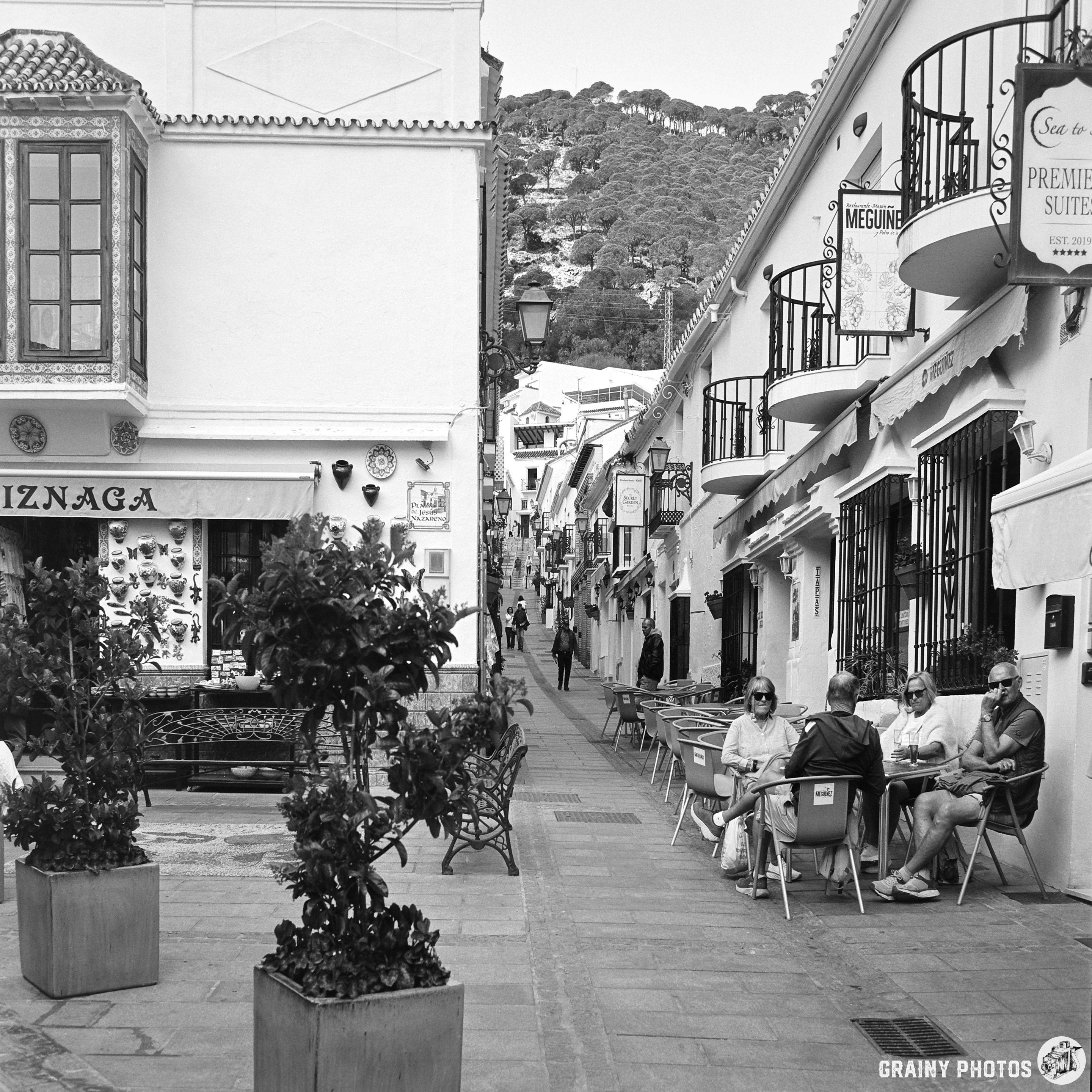 A charming narrow street in Mijas, lined with white buildings featuring balconies. People sit at outdoor cafes, enjoying the atmosphere. Potted plants and local shops add to the rustic charm of this scenic destination.