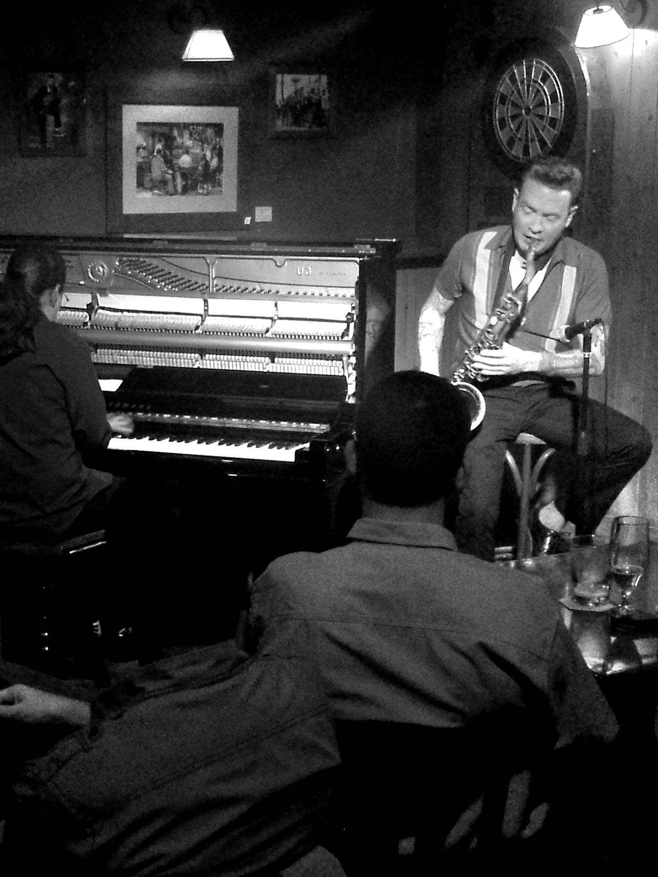 The black and white, vertical photograph was taken in 2014 at the now-closed Honky Tonk Blues Bar in Barcelona. It shows musicians Lluís Coloma—with his back to the camera, playing an open upright piano—and Dani Nel·lo, seated, playing the saxophone. The bar's decor is visible, and in the foreground, the silhouettes of spectators seated at their tables with drinks.